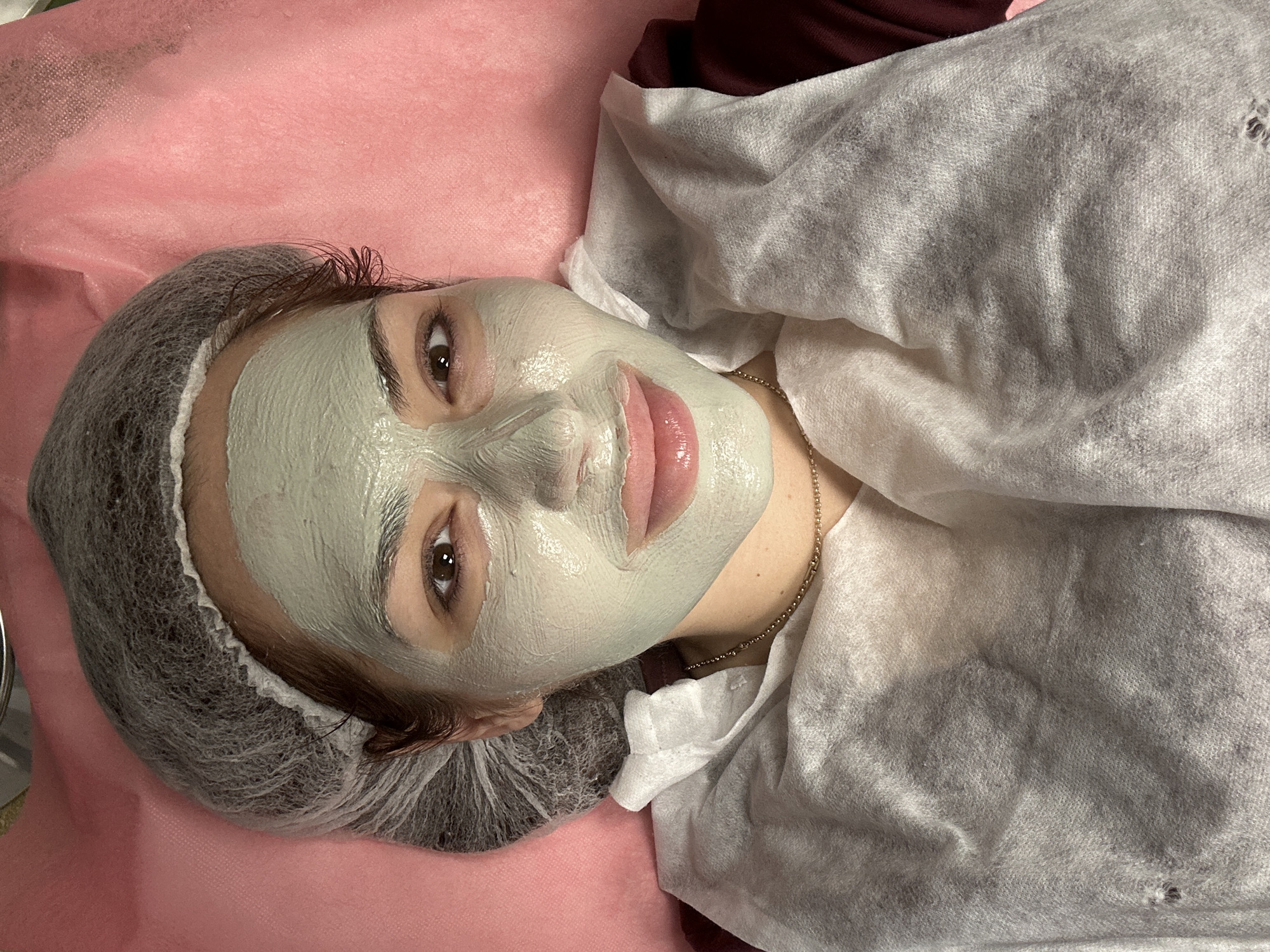A woman lies on a pink surface, covered by a white, porous material, with a gray hair net covering her hair. A light green mask covers her face. A gold chain rests around her neck. The background is dark. The location cannot be determined based on the image provided.
