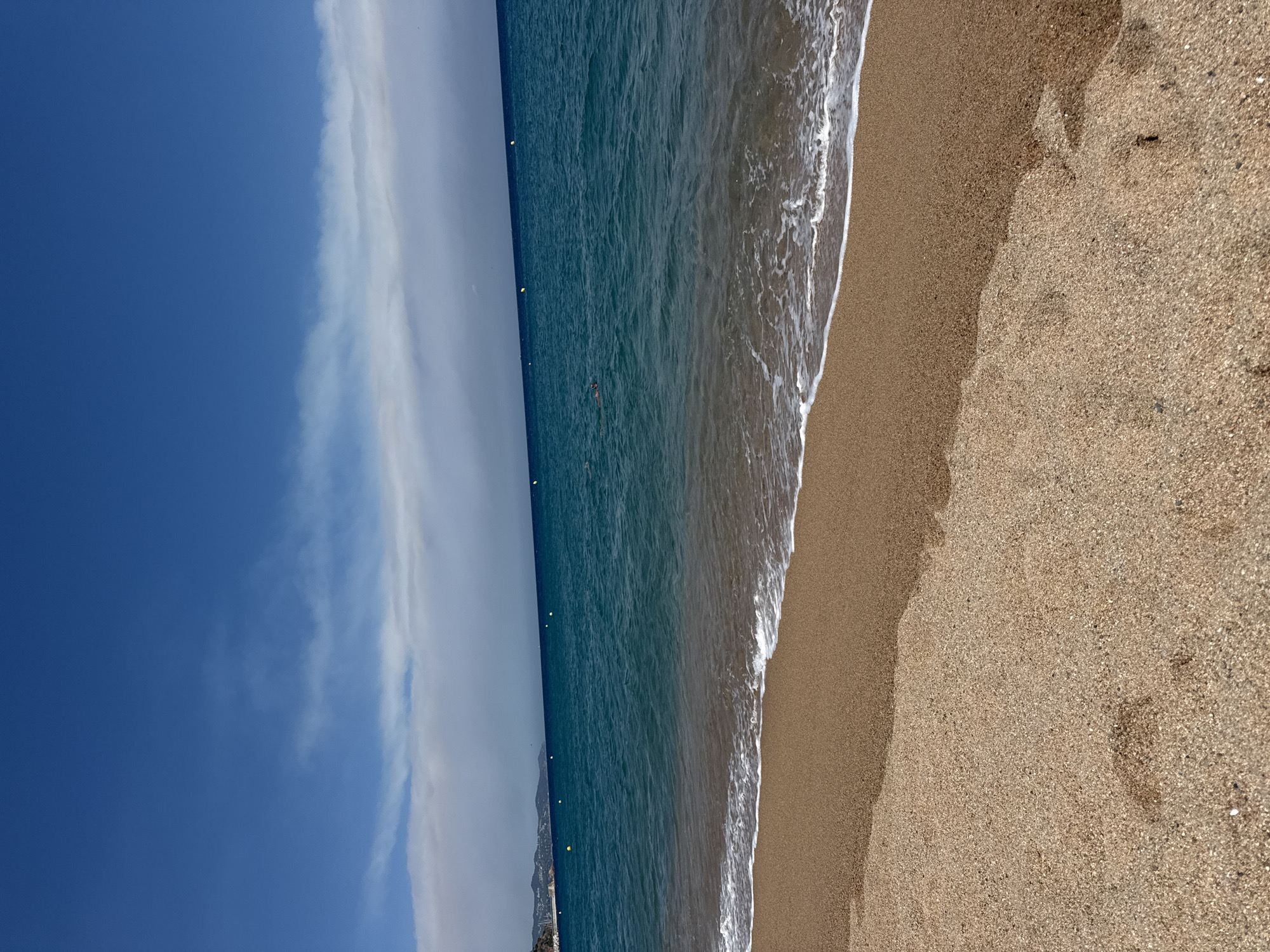 The image depicts a sandy beach in Blanes, Spain, with the turquoise Mediterranean Sea stretching towards the horizon. Gentle waves lap at the shore, leaving a thin white foam line on the wet sand. The sky is a vibrant blue, with a band of wispy white clouds in the distance. In the far distance, a coastline with some buildings and hills is visible. Several yellow buoys are scattered in the sea. In the middle ground of the sea, two individuals are visible swimming or snorkeling. The foreground is dominated by the textured, light brown sand, with some footprints visible. The overall scene is tranquil and suggests a sunny day at the beach.