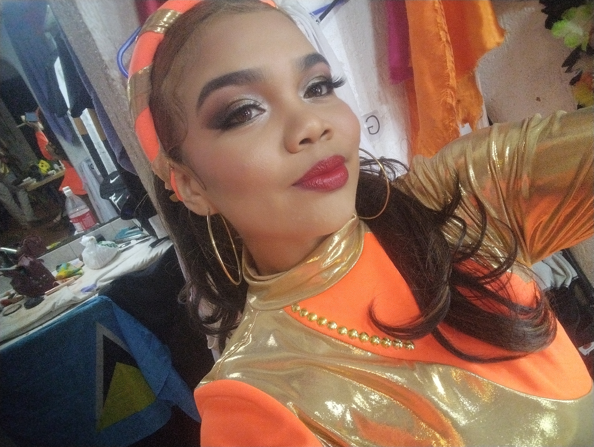 The image depicts a woman in an orange and gold shiny outfit with a matching headband and large hoop earrings. She has dark hair, brown eyes, and is wearing elaborate makeup including dark eyeshadow, false eyelashes, and red lipstick. The background appears to be a cluttered room with clothing racks, a table with various items on it, and a blue object with a yellow triangle design. Based on the Saint Lucian flag visible, she may be of Saint Lucian heritage, or it may be related to the costume/performance. Without additional information, I cannot confirm the exact location in Porlamar, Venezuela.