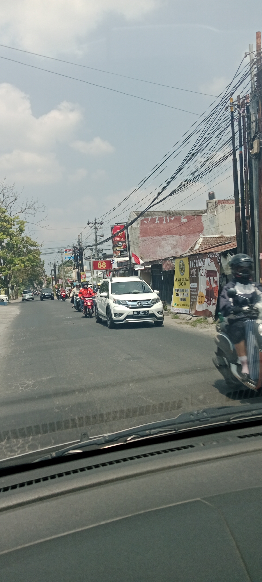A street view shows a traffic jam on a sunny day in Trihanggo, Indonesia. A white SUV, license plate AB 1322 XH, is in the foreground, followed by a line of motorcycles and other vehicles. Several motorcyclists wear helmets. One motorcyclist in a red jacket is prominent. Buildings line the right side of the street, with advertisements and overhead power lines. Trees and more buildings are visible on the left. The perspective is from inside a vehicle, looking through the windshield, which shows the dashboard.