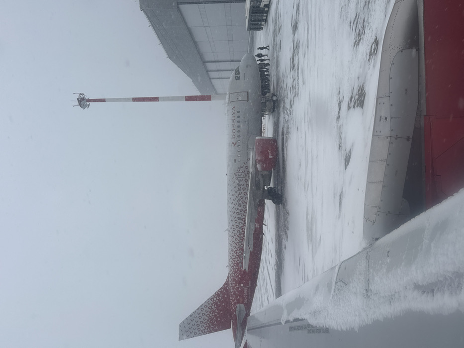 The image captures an airport apron at Емельяново (аэропорт), Russia, during an ongoing snowfall. A Rossiya Airlines Airbus aircraft, registration RA-73190, is stationary on the snow-covered tarmac. The aircraft features a light gray fuselage with a distinctive red and white patterned livery on its tail and engine nacelles, and "ROSSIYA" lettering on its side. Approximately 10 to 15 individuals, dressed in dark winter clothing, are congregated near the front right main cabin door of the aircraft, with some appearing to move towards the right side of the frame. To the right of the aircraft, two white airport buses are partially visible. In the background, a tall red and white striped mast or communication tower stands adjacent to a large, light-colored building, likely a hangar or terminal, which extends across the right side of the background. The foreground reveals a snow-covered aircraft wing and a portion of a red engine cowling, indicating the image was taken from another parked aircraft. The entire scene is under overcast skies, with visible snowflakes falling and accumulated snow covering all exposed surfaces.