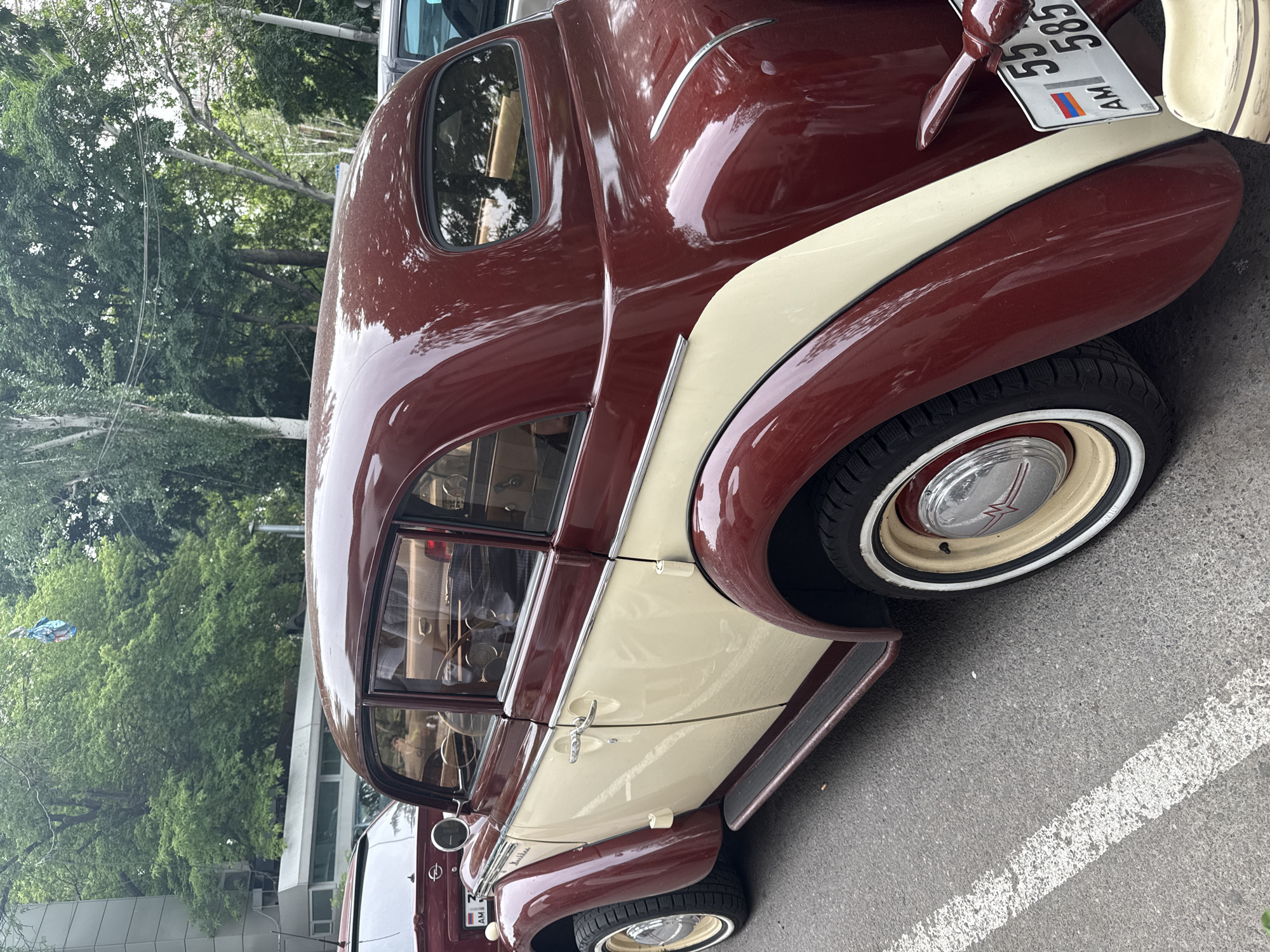 The image shows a vintage maroon and cream-colored car parked on an asphalt surface. The car has a license plate that reads "55 SS 585 AM," which indicates that the car is registered in Armenia. The background includes trees and buildings, suggesting an urban setting, likely in Yerevan, Armenia. The car is parked near a white parking line. The tire has a white wall. The car's chrome trim and polished paint are reflective.