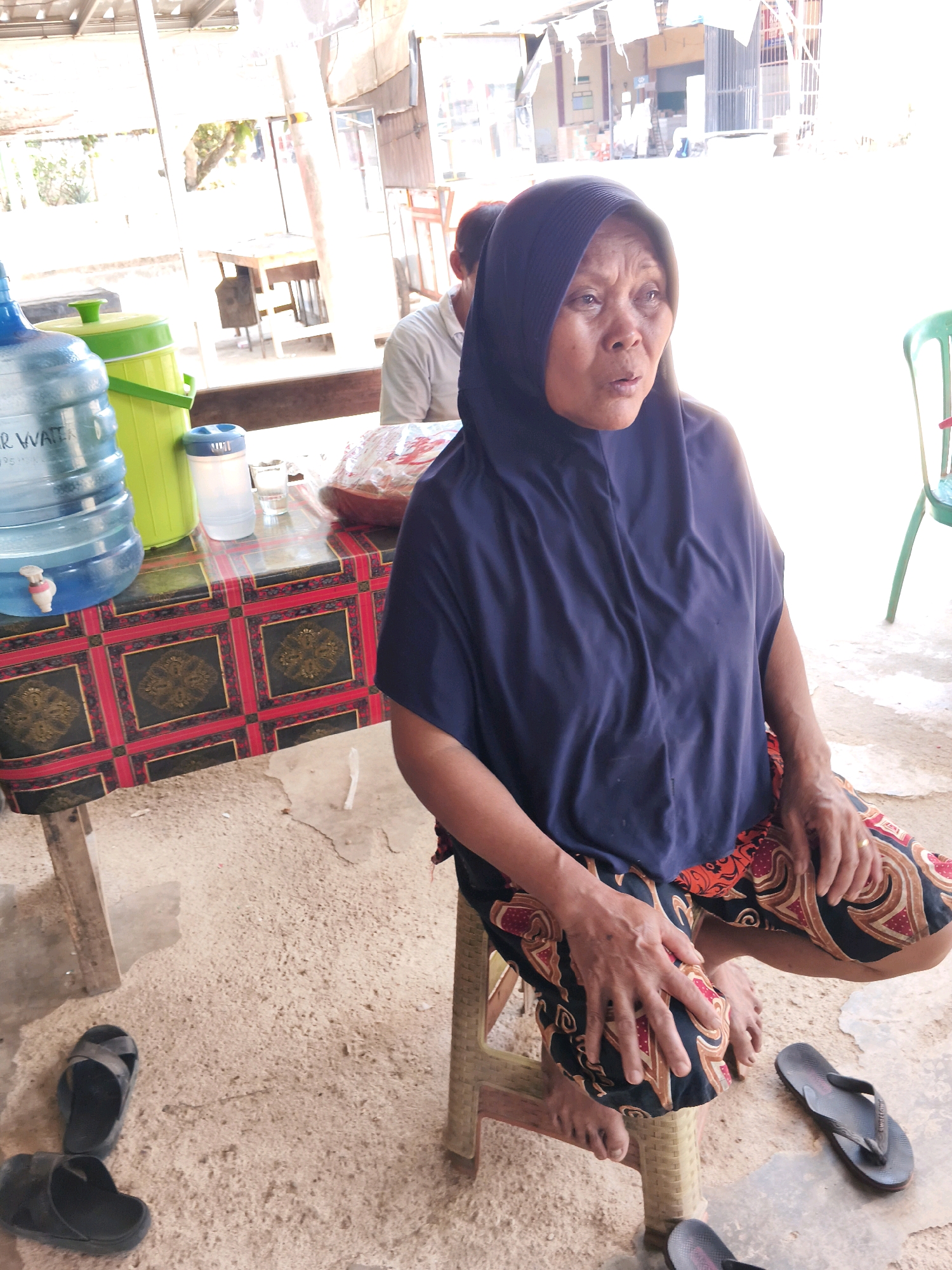The image depicts an elderly Indonesian woman wearing a dark blue hijab and a patterned sarong, seated on a light brown plastic stool. She is positioned in what appears to be an outdoor or semi-outdoor setting in Sendang Retno, Indonesia. Her gaze is directed to her right, and her mouth is slightly open, suggesting she may be speaking or in mid-action. Her hands rest on her lap, with a ring visible on her left hand.
To the left of the woman, a table is covered with a red and black patterned cloth. On the table, there is a large blue water jug with a white spigot, a green cooler, a white plastic bottle with a blue lid, and a glass of water. In the background, a person wearing a light-colored shirt is partially visible.
The ground is sandy and somewhat uneven. Several pairs of black flip-flops are scattered around the area, with two pairs prominently visible in the foreground. The environment suggests a casual, possibly roadside or market setting, characterized by simple structures and the presence of everyday items. The lighting indicates it is daytime, with sunlight casting shadows.