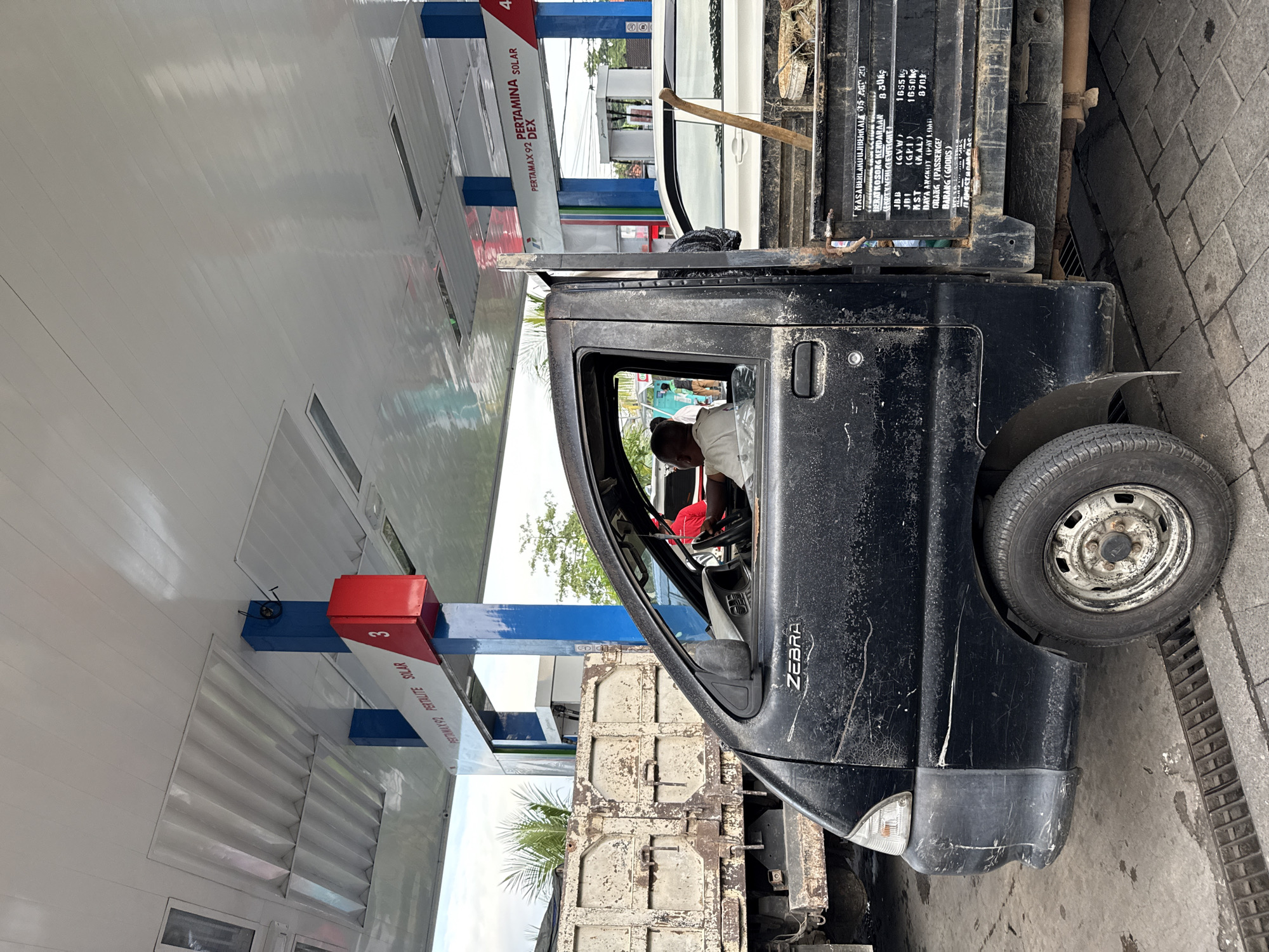A black, weathered "Zebra" pickup truck is parked at a gas station in Ungasan, Indonesia. The driver, wearing a white shirt, is visible inside the vehicle. The truck is positioned beside a gas pump displaying "Pertamina" signage for "Pertamax 92," "Pertamax Solar," and "Dex." Text on the truck's cargo area includes weight and load capacity information. The gas station's canopy is visible overhead. The ground is paved with cobblestones.