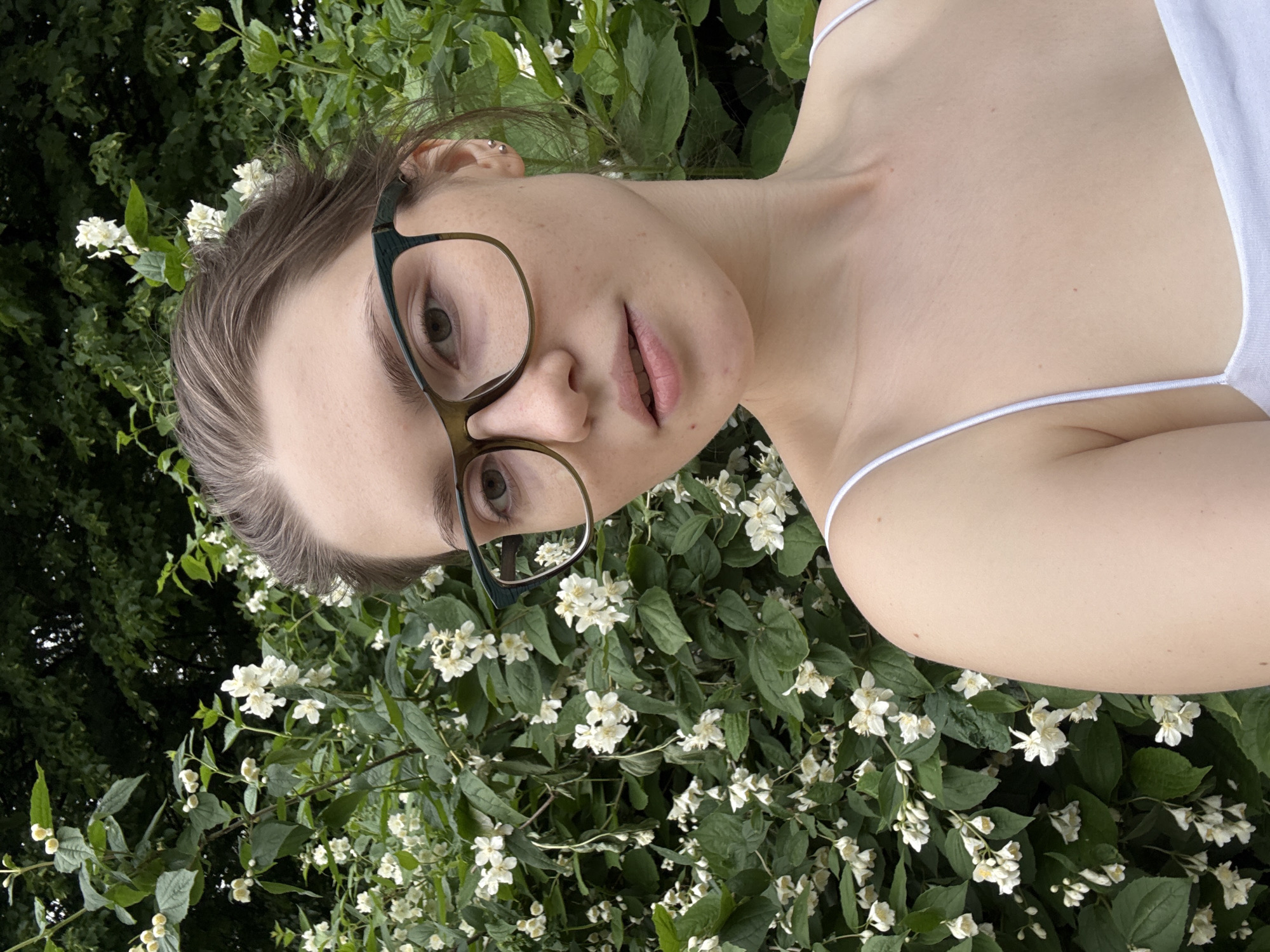 A young woman with brown hair pulled back is looking directly at the camera. She is wearing glasses with thick, dark green frames and a white tank top. Her skin is fair, and she has subtle freckles on her cheeks. The background consists of lush green foliage and small white flowers, likely a blooming bush. The lighting suggests it is daytime, and the overall setting appears to be outdoors, possibly in a park or garden. The image was taken in Saint Petersburg, Russia.