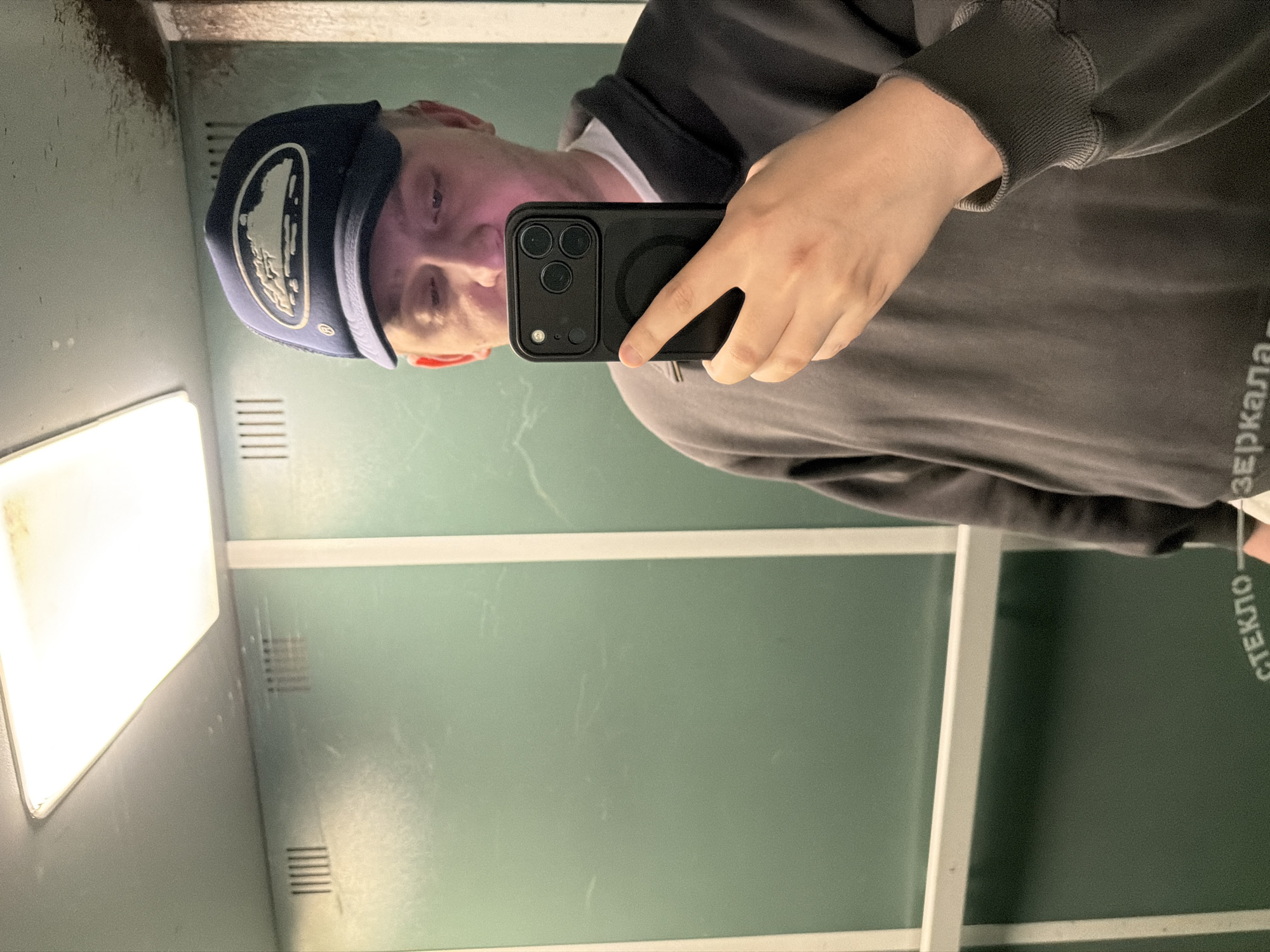 A person wearing a dark blue baseball cap and a dark gray sweatshirt is taking a selfie in an elevator. The elevator has green walls, a white ceiling light, and a metallic vent on the wall. The person is holding a black smartphone in their right hand, which is pointed towards a mirror, capturing their reflection. The elevator also appears to have some text on the lower part of the wall, but it is partially obscured. The location is identified as an elevator in the city of Khimki, Russia.