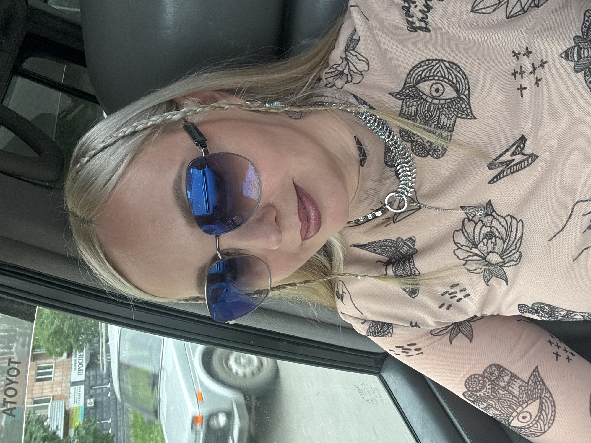 A woman with blonde hair in braids is sitting in the passenger seat of a car. She is wearing sunglasses and a light-colored shirt with black line drawings of hands, eyes, flowers, and butterflies. A silver chain necklace is also visible. Through the car window, a light-colored car and a building with the signs "AToyot" and "Проспект" can be seen. The location is Komsomolsk-on-Amur, Russia.