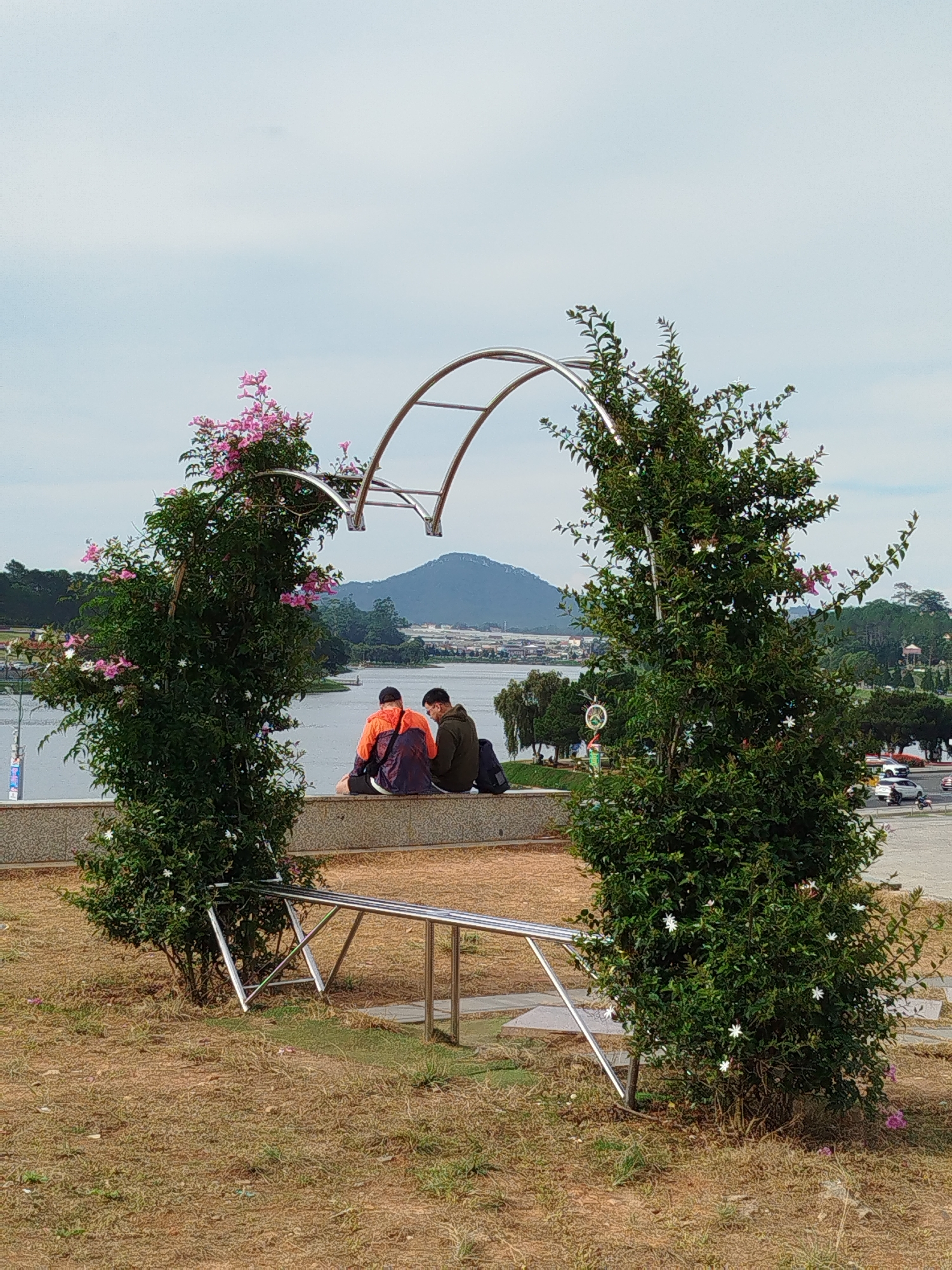 Two individuals are seated on a retaining wall, observed through a decorative archway adorned with foliage and flowers. The archway, constructed of stainless steel, frames the pair and the landscape beyond. The person on the left is wearing an orange jacket, while the person on the right is in a green jacket. The background features a body of water, presumably a lake, with a mountain visible in the distance. The sky is overcast. A few cars can be seen on the right side of the frame. The location is identified as Da Lat, Vietnam, based on the architectural and environmental characteristics.