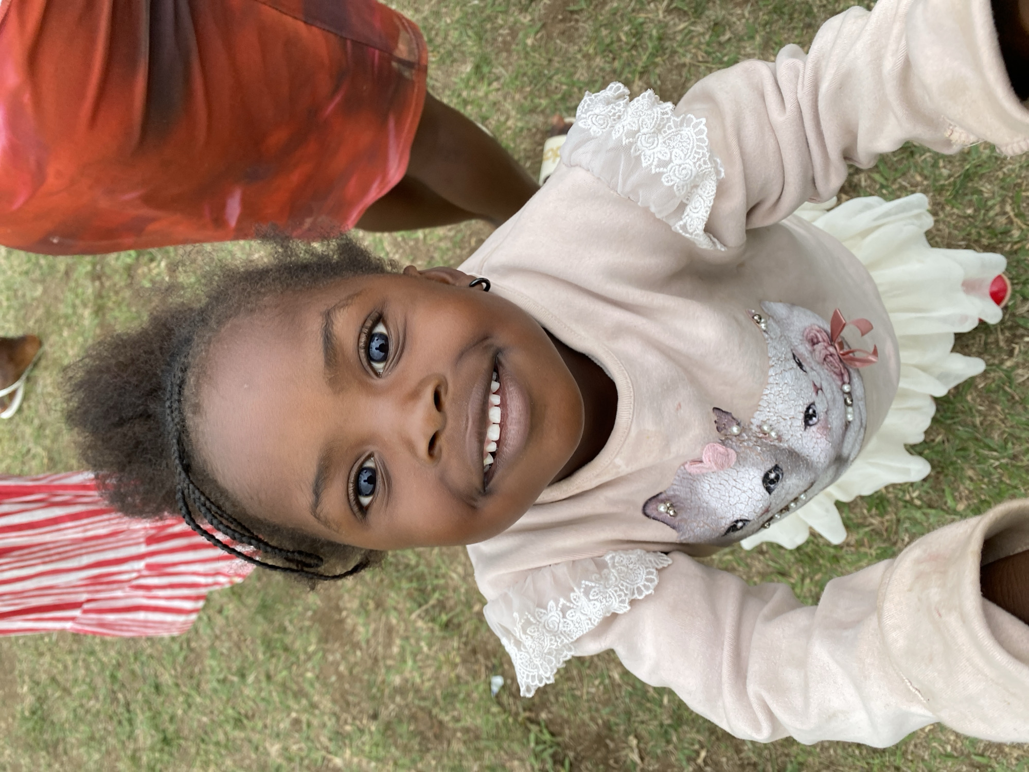 A young girl with dark skin and braided hair is smiling and looking up at the camera. She is wearing a light pink long-sleeved shirt with white lace details and a white skirt. The background shows green grass and parts of other people's clothing, suggesting an outdoor location. The image was taken in Kuru Dungus, Nigeria.