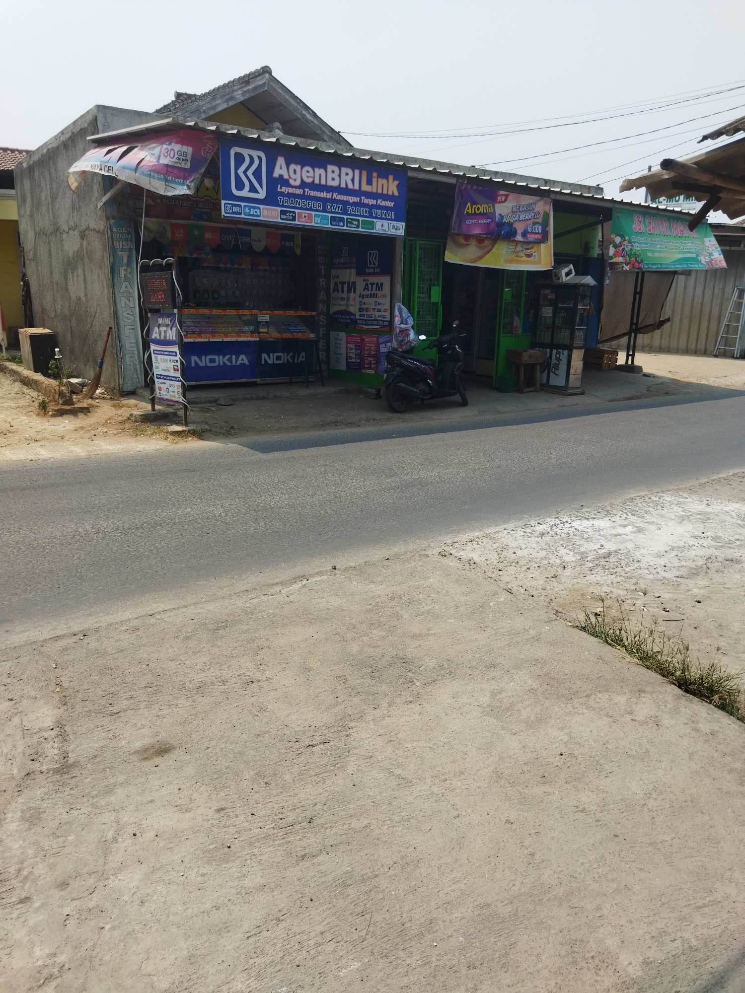 The image shows a street view of a small shop in Suka Sari, Indonesia. The shop appears to be a local convenience store or service center, with signs indicating services like "AgenBRI Link" which offers financial transactions, and "JISI SAYUR MAYUR" selling vegetables. There are advertisements for mobile phone data plans and various brands. A motorcycle is parked in front of the shop. The building is a single-story structure with a corrugated metal roof. The street is paved and the area is lined with concrete sidewalks. The overall scene depicts a typical street scene in a residential or commercial area of Indonesia.