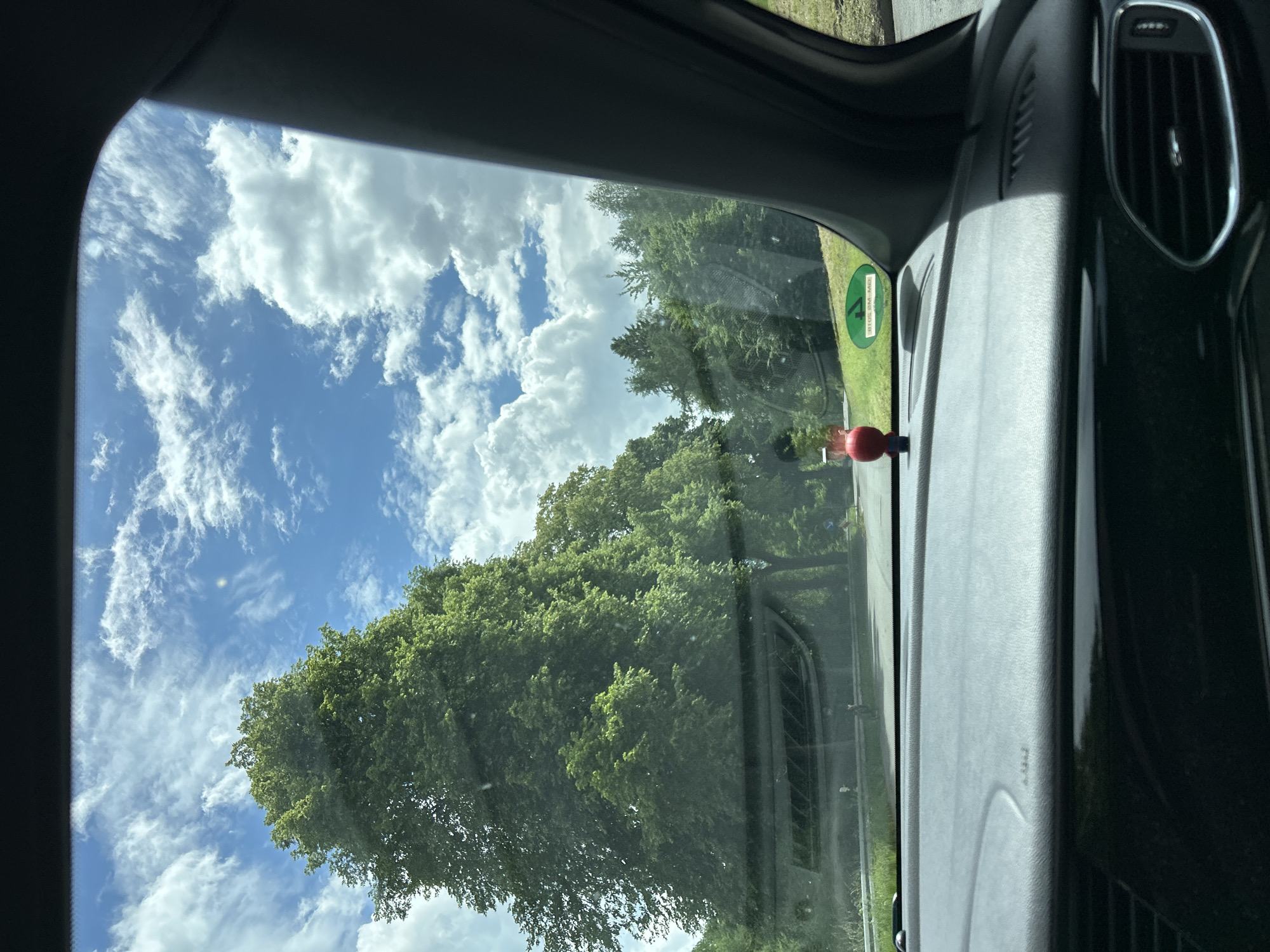 The image presents a forward-facing view from inside a vehicle, observed through the windshield, on a clear day in Reichshof, Germany. The sky occupies the upper portion of the frame, displaying a vibrant blue with scattered, white cumulus clouds. The sun appears to be high, indicated by bright illumination and a faint lens flare visible in the upper right quadrant of the sky.
The immediate foreground reveals the vehicle's dark gray dashboard, with the word "AIRBAG" faintly embossed on the passenger side. A black ventilation grille is visible on the far right of the dashboard. On the dashboard, slightly left of center, a small red spherical object, possibly a figurine or ornament, is positioned. Adjacent to the right A-pillar, a green circular sticker is affixed to the windshield. The sticker displays the number "4" prominently in its center, with "30105" inscribed at the top and "KM-MD" at the bottom, characteristic of a German environmental badge. The dark gray interior of the car's right A-pillar is visible along the right edge of the frame.
Beyond the windshield, an asphalt road curves gently to the right. A silver-colored metal guardrail runs along the left side of the road. Dense green foliage dominates the midground and background. On the left, large deciduous trees with full, green canopies line the road. On the right, a mix of coniferous and deciduous trees forms a dense forest alongside the road. A small blue sign with white pictograms is faintly discernible in the distance on the right side of the road, partially obscured by the trees. No individuals or animals are visible in the scene. The overall environment suggests a natural, possibly rural or semi-rural, setting.