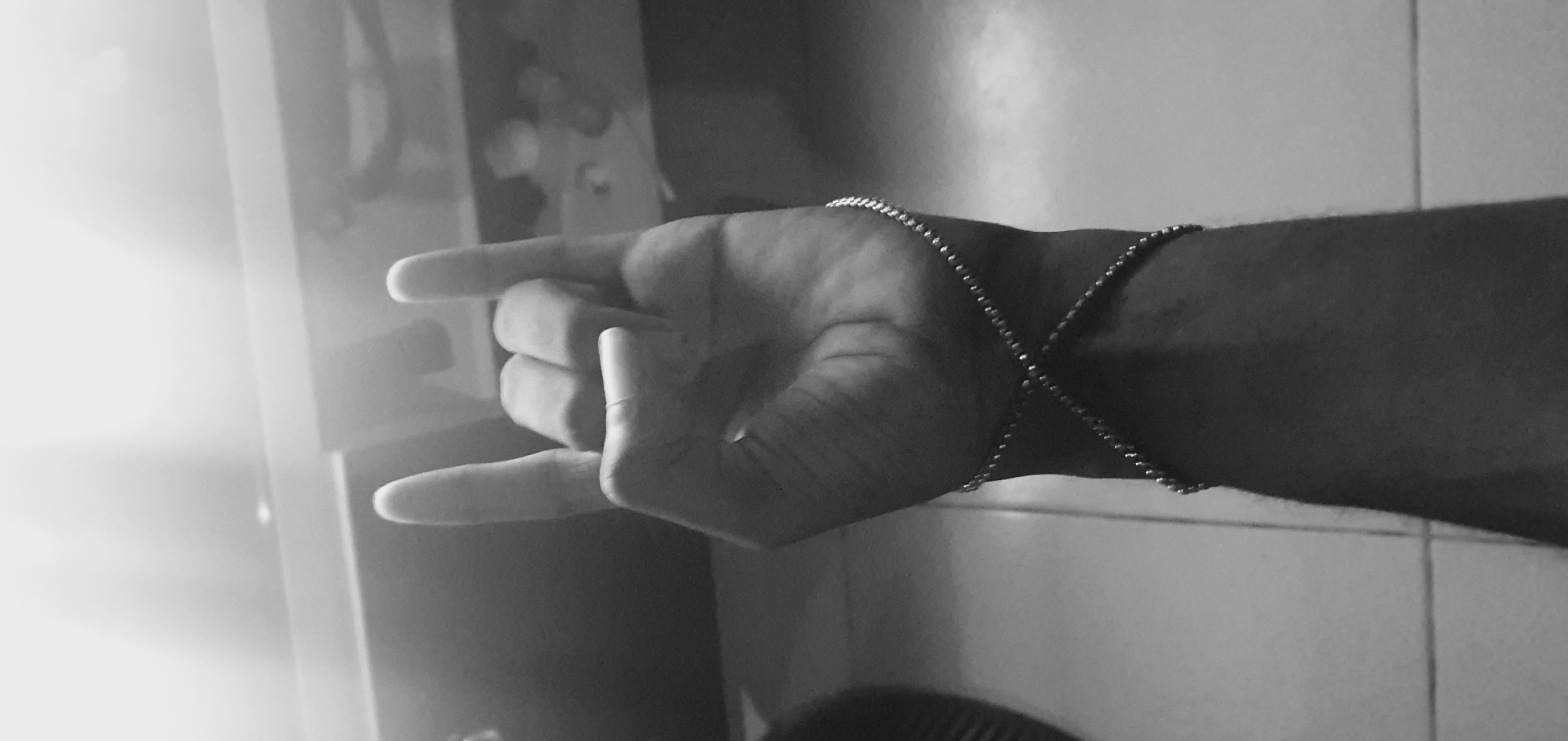 A black and white image shows a person's hand making a "rock on" gesture. A thin chain bracelet is visible on the wrist, crisscrossing the forearm. The background appears to be a room with tiled walls. The image does not contain sufficient information to determine a specific location within Lokoja, Nigeria.