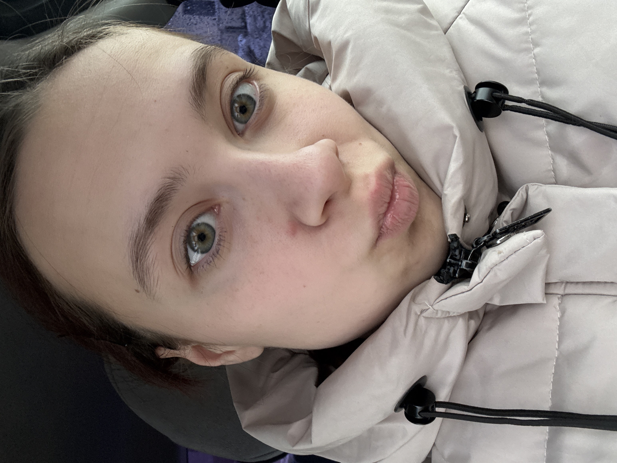 A close-up, angled portrait shows a young woman with fair skin and light brown eyebrows and hair, wearing a light beige puffer jacket. Her eyes are green, and she has a mole on her cheek. She is making a kissing face with slightly parted lips. The jacket has black toggles and drawstrings visible. The location of the image is not determinable from the provided image.
