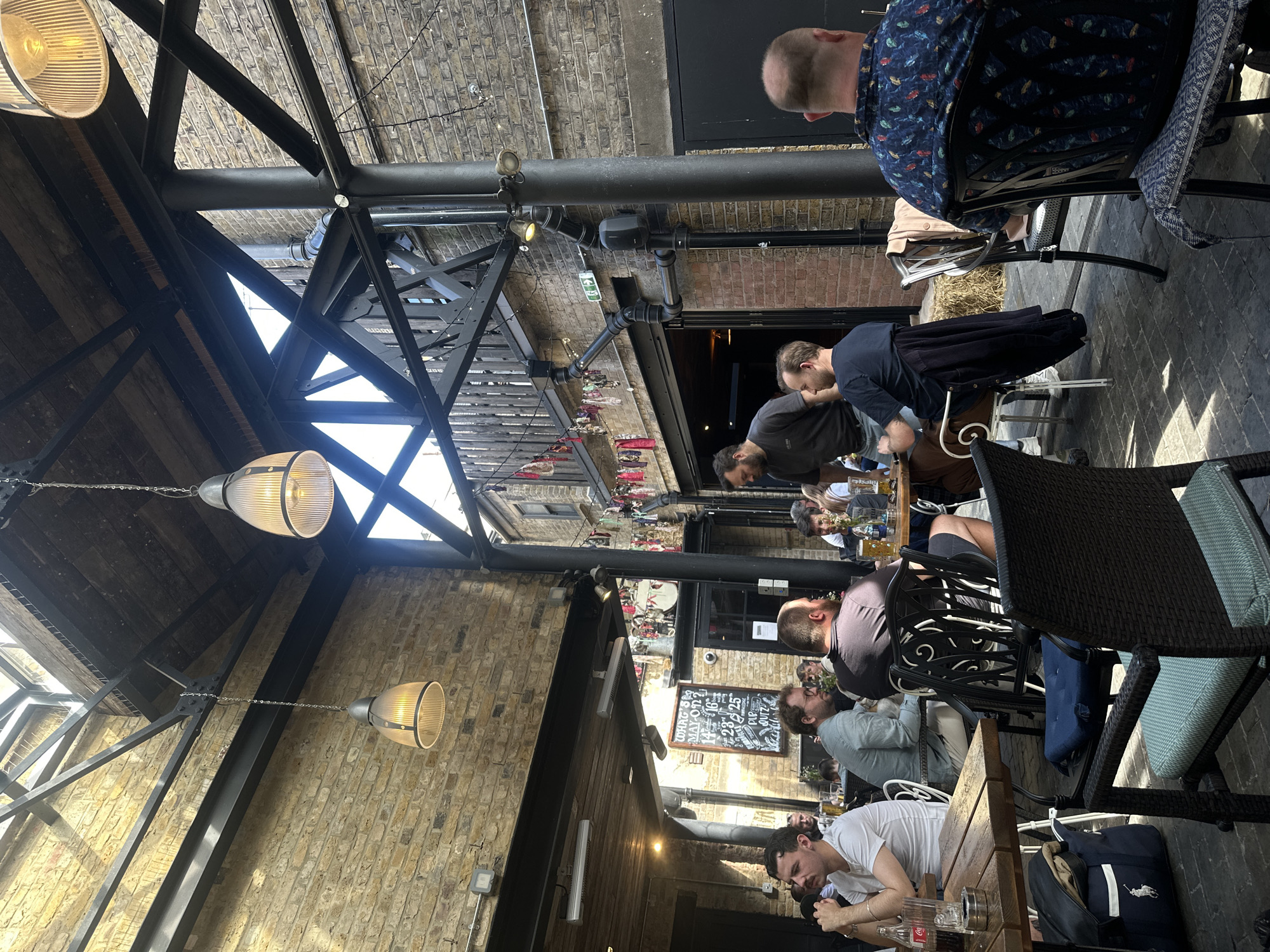 The image shows an outdoor seating area of a pub or restaurant in London, United Kingdom. The architecture is industrial, with exposed brick walls, black metal beams supporting a partial roof, and pendant lights. Several people are seated at tables, some engaged in conversation or looking at their phones. The tables are wooden, and the chairs are a mix of wicker and metal. There's a chalkboard menu visible on the wall. The scene is well-lit, suggesting it's daytime.