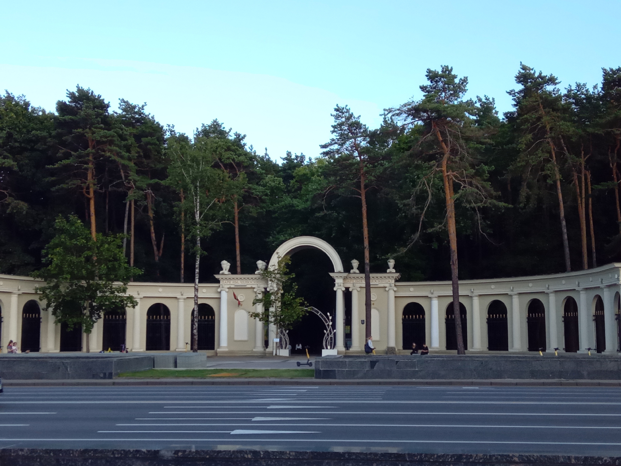 The image shows a wide view of an architectural structure with a series of arches and columns, reminiscent of a colonnade or a grand entrance. The structure is light-colored, likely stone or stucco, and curved, extending across the frame. Behind the structure, a dense forest of tall, slender trees, predominantly pines, forms a dark green backdrop. The sky above is a clear, pale blue, indicating daytime, possibly late afternoon or early evening.
In the foreground, a multi-lane asphalt road runs horizontally, with white lane markings clearly visible. A dark, textured border is present at the very bottom of the image, possibly a curb or a wall.
In the middle ground, several elements suggest a public park or recreational area. There is a stone or concrete barrier in front of the colonnade. A few people are visible near the colonnade, some sitting on the barrier and others standing. There are also some yellow electric scooters parked near the structure.
The architectural centerpiece of the scene is a large, arched gateway in the center, framed by columns. Above the arches, there are decorative elements, including urn-like sculptures on pedestals. Text is visible above some of the arches, although it is mostly illegible due to the distance and resolution. One snippet appears to read "ПАРК ИМ. ЧЕЛЮСКИНЦЕВ" (Park named after Chelyuskinets), indicating the location is the Chelyuskinets Park in Minsk, Belarus.
The overall impression is a blend of grand classical architecture and the natural beauty of a dense forest. The presence of people and scooters suggests the park is active and frequented by visitors. The lighting indicates a relatively calm, perhaps late-day atmosphere.