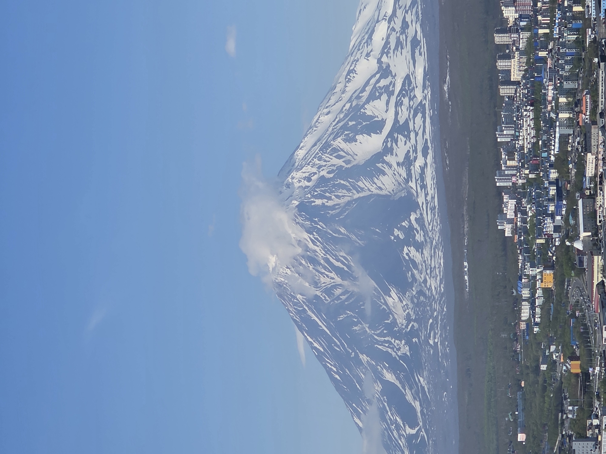 A wide shot captures a snow-capped volcano dominating the background, its peak shrouded in a wispy cloud. Below the volcano, a dense forest separates it from a sprawling city. The city is a mosaic of low-rise and mid-rise buildings, their rooftops a mix of colors. Roads weave through the urban landscape, with cars visible on some of them. The sky is a clear, vibrant blue, with a few faint clouds scattered about. The overall impression is of a city nestled at the foot of a majestic natural wonder.
