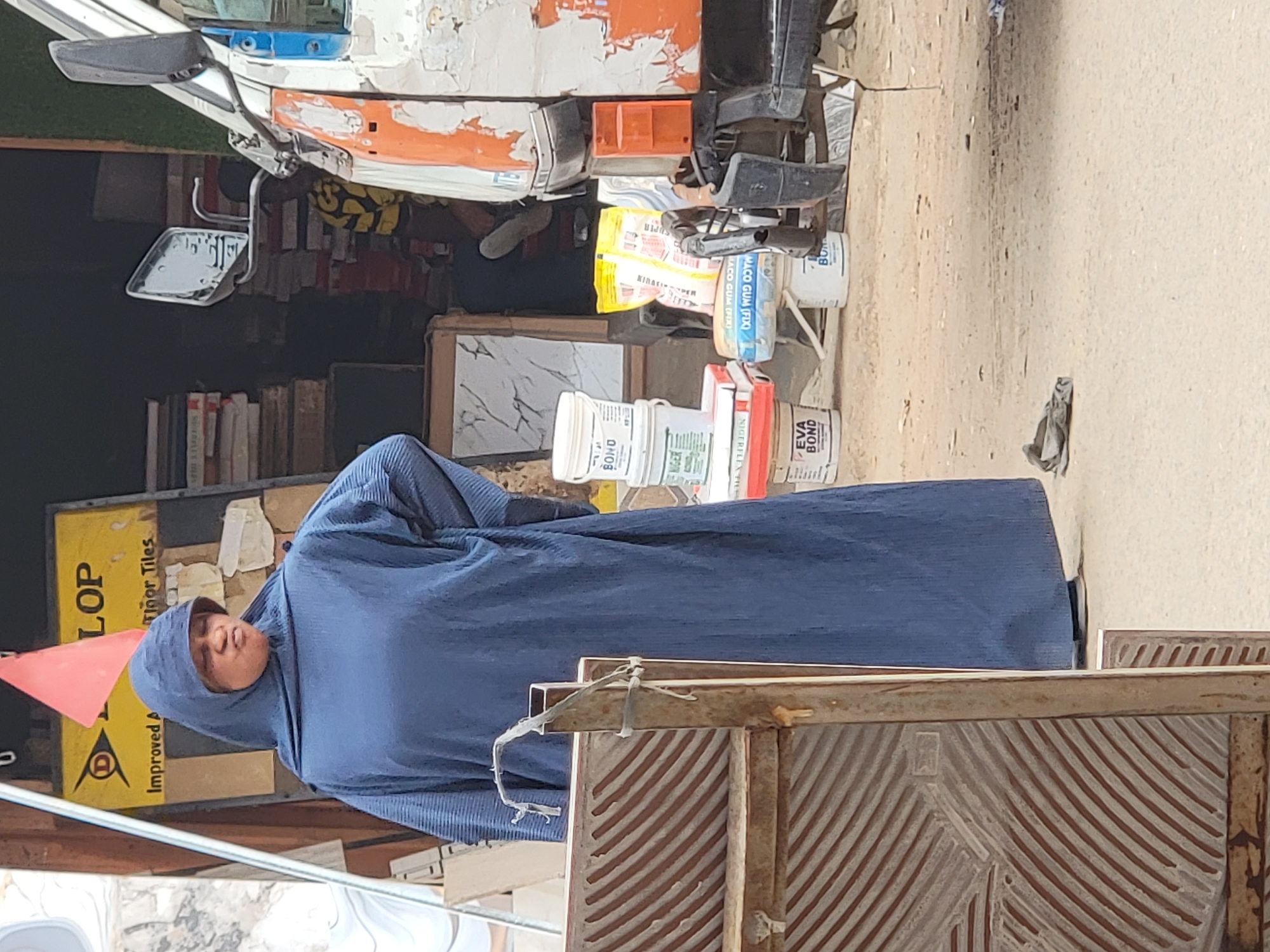 This image captures an outdoor scene in Maiburiji, Nigeria, likely a street or market stall. In the foreground, a person wearing a full-length blue garment and matching hijab stands, facing slightly to their left.
Behind the person, to the left, is a display of various goods. A white and orange vehicle, possibly a small truck or tricycle taxi, is partially visible at the top left, showing a side mirror and part of its bodywork. Below the vehicle, shelves stocked with books are present. On the ground and stacked near the stall are numerous white plastic buckets and containers. Visible text on these containers includes "BOND," "NIGERFLEX," "NIGER," "EVA BOND," and "MACO GUM (FIX)," suggesting they contain adhesives or other building materials. A patterned tile sample is also visible among these items. A yellow sign partially reads "LOP," "Improved A," and "Floor Tiles," further indicating a tile or construction material vendor.
The ground is unpaved, appearing as dirt or sand. A piece of crumpled fabric or trash lies on the ground to the right. In the immediate foreground on the right, a rustic gate or fence structure, made of wood and metal, is partially visible. The scene is brightly lit, indicating daytime and clear weather conditions. The person appears to be observing or passing through the area.