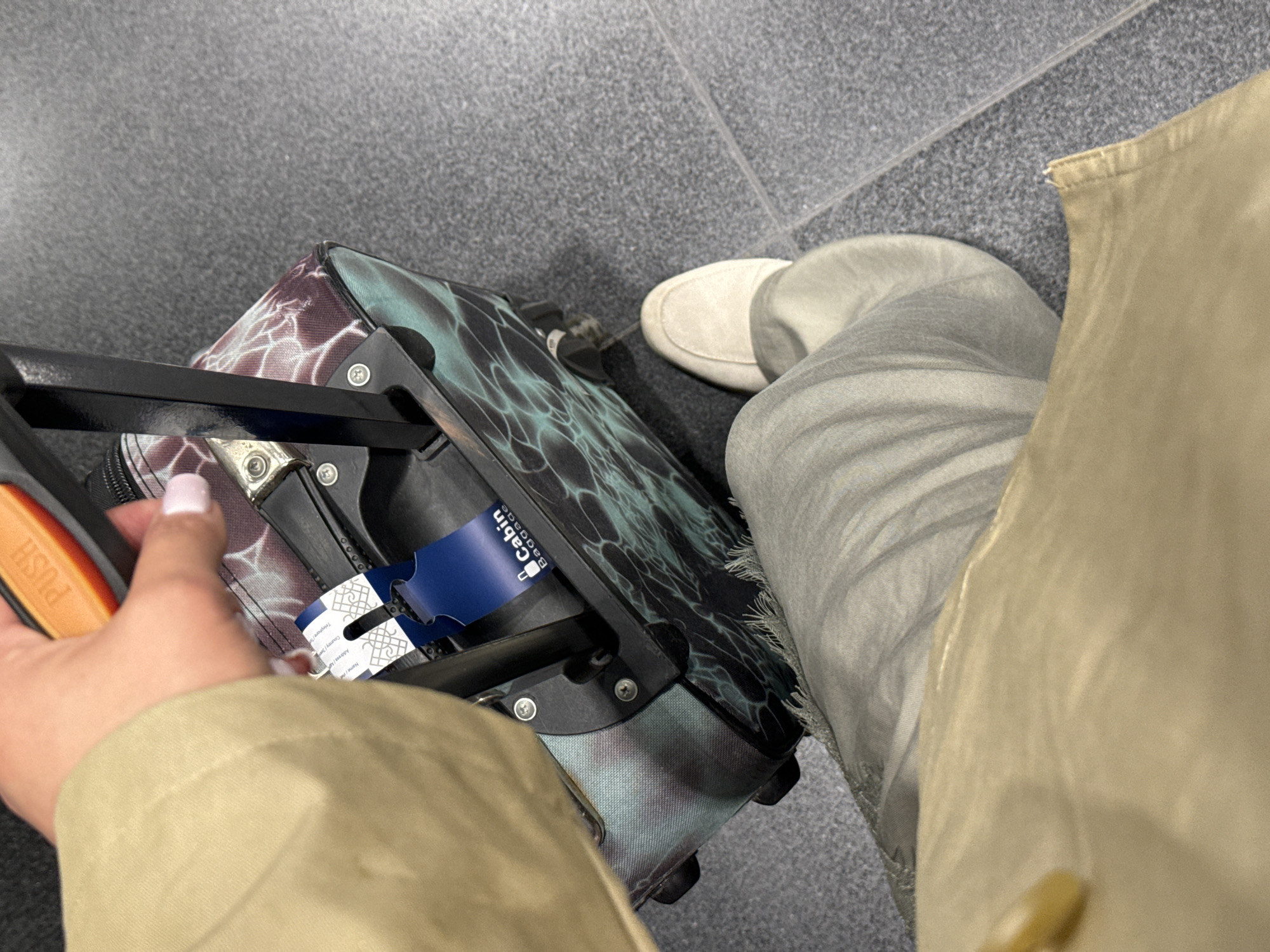 A person stands on gray, tiled flooring in what appears to be an airport or travel hub. They are wearing gray slacks and a beige shoe, and a light brown jacket. To their left is a colorful carry-on suitcase with a black handle, equipped with a luggage tag labeled "Cabin Baggage." The person's left hand is visible holding the handle, with neatly manicured fingernails. The background suggests an indoor, possibly transit-related environment. The location in Khimki, Russia, is not directly evident from the visual details in the image.
