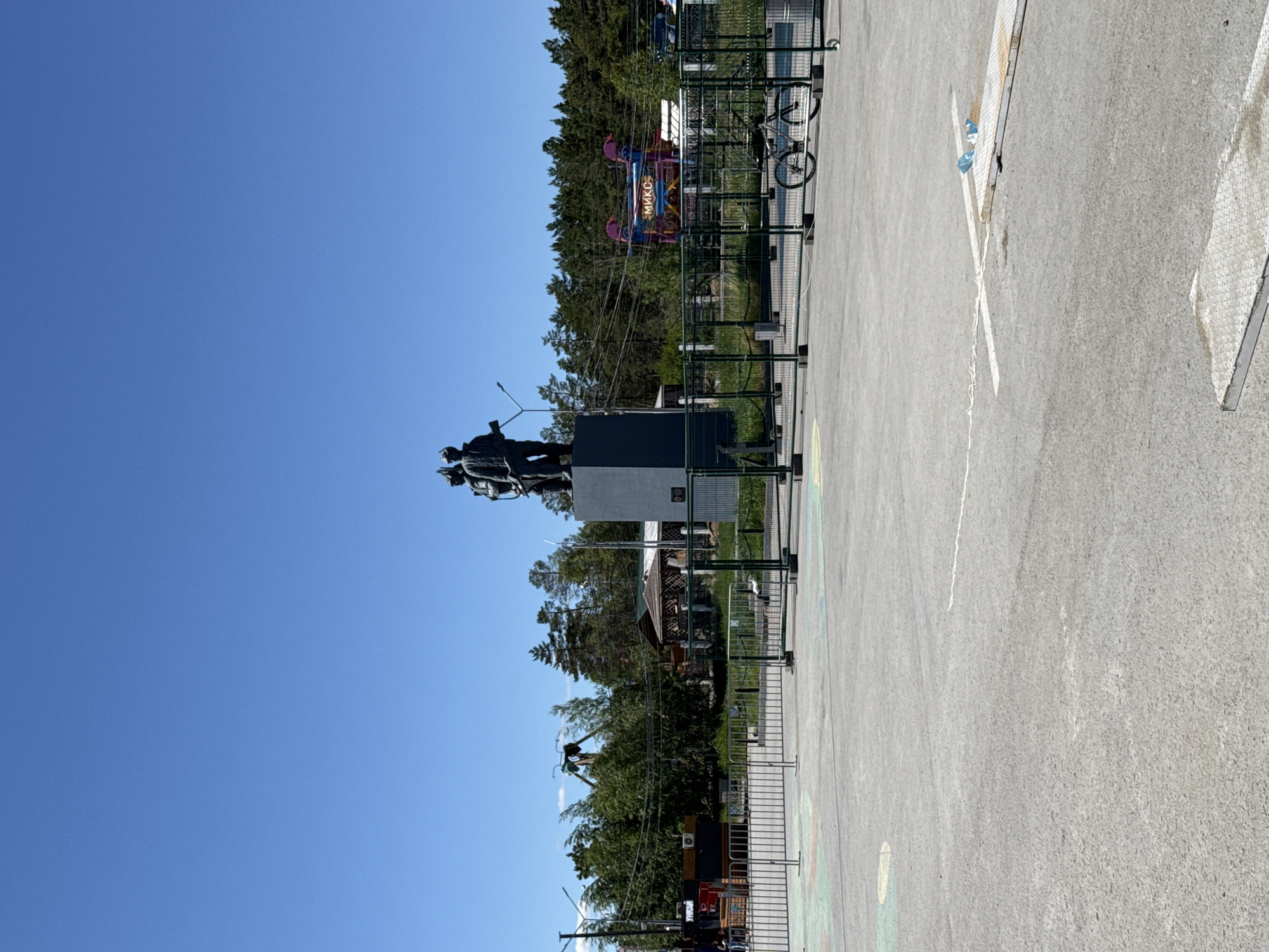 In Yakutsk, Russia, a wide, paved plaza is the foreground of this image, marked with painted lines and some debris. A metal fence separates the plaza from the background. Behind the fence, several amusement park rides are visible amidst trees. To the right, a bicycle is parked next to the fence. In the center of the background, a dark gray monument depicting three figures stands on a pedestal. Beyond the monument, a vibrant amusement ride with the word "МИКС" (MIX) is visible. The sky is clear and blue.