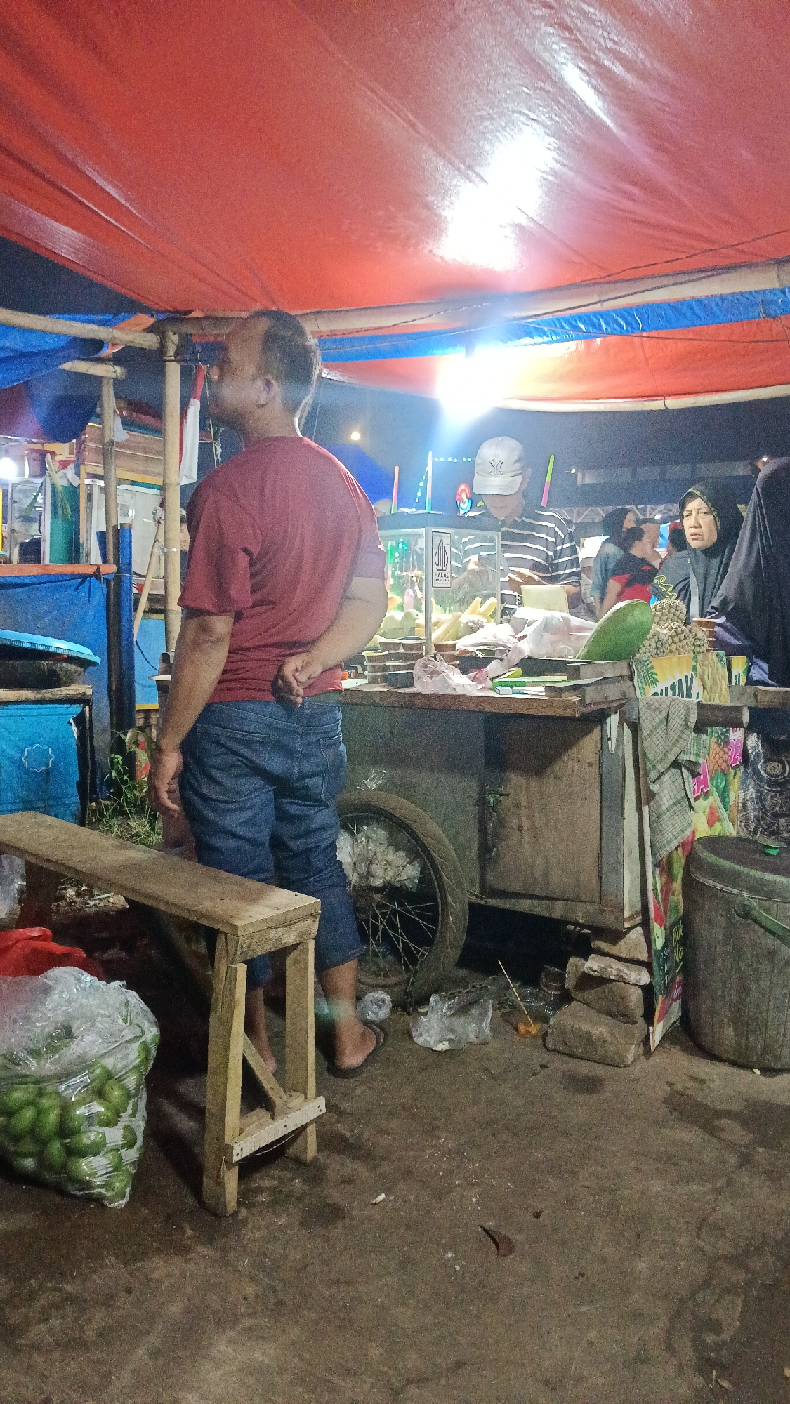 In Tajurhalang, Indonesia, a street food vendor operates under a makeshift canopy of red and blue tarps. A man in a maroon t-shirt and blue jeans stands with his back to the camera, his hands clasped behind him. He appears to be a customer or bystander. In the background, a vendor wearing a striped shirt and a white baseball cap serves customers from a cart filled with fruits and what appears to be juice dispensers. Two women, one in a black hijab and the other partially visible, are also present at the stall. A large, clear plastic bag filled with green fruits, possibly avocados, sits on the ground near a wooden bench. The ground is damp and uneven, typical of an outdoor market environment. A bicycle wheel is attached to the vendor's cart. The scene is illuminated by overhead lighting, casting a warm glow on the surroundings.