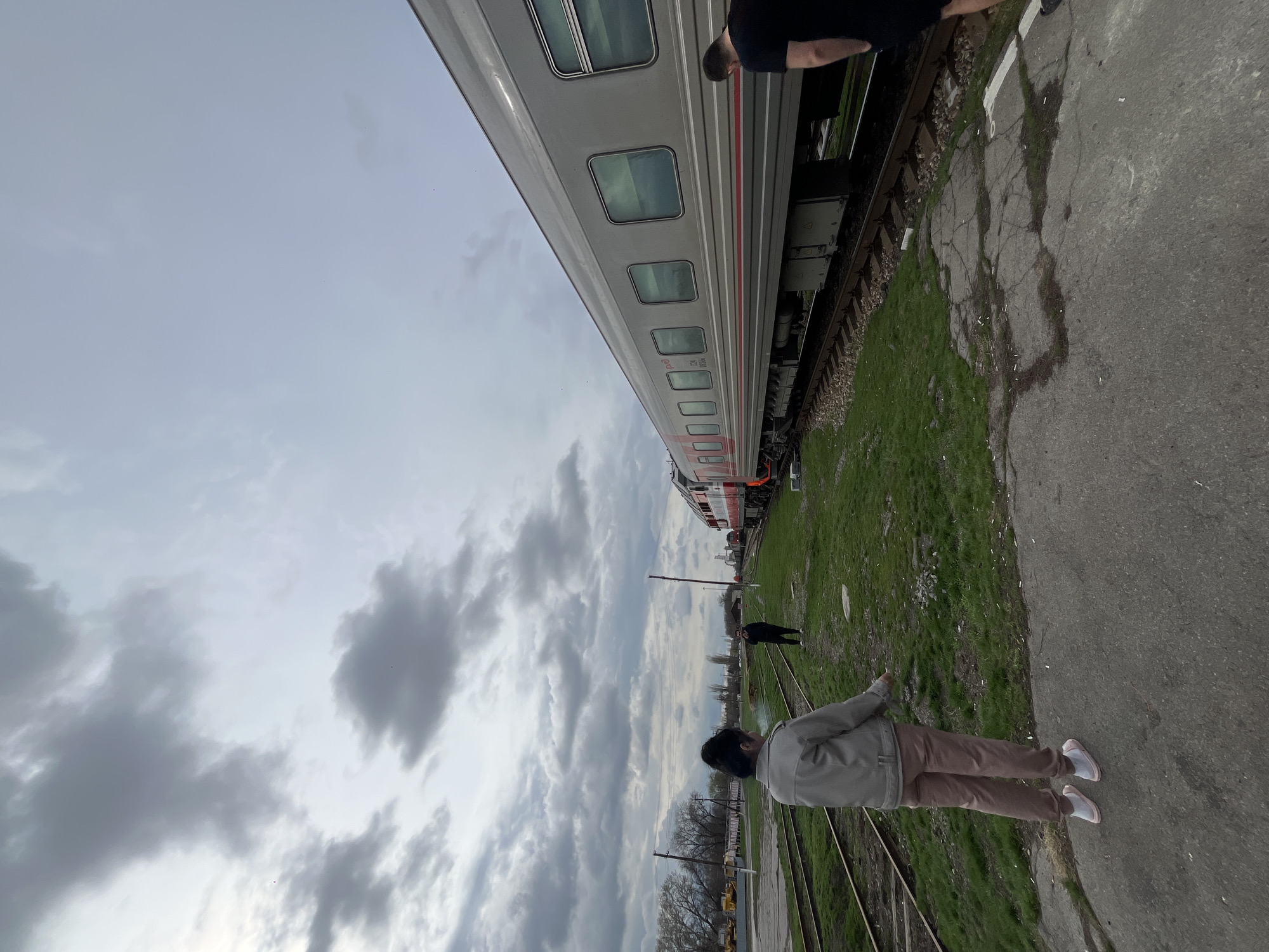 The image shows a train parked next to a cracked concrete platform and grassy area in Morozovsk, Russia. A person stands on the platform facing away from the camera. Another person is further down the platform, closer to the train. Trees are visible in the background. The sky is cloudy.