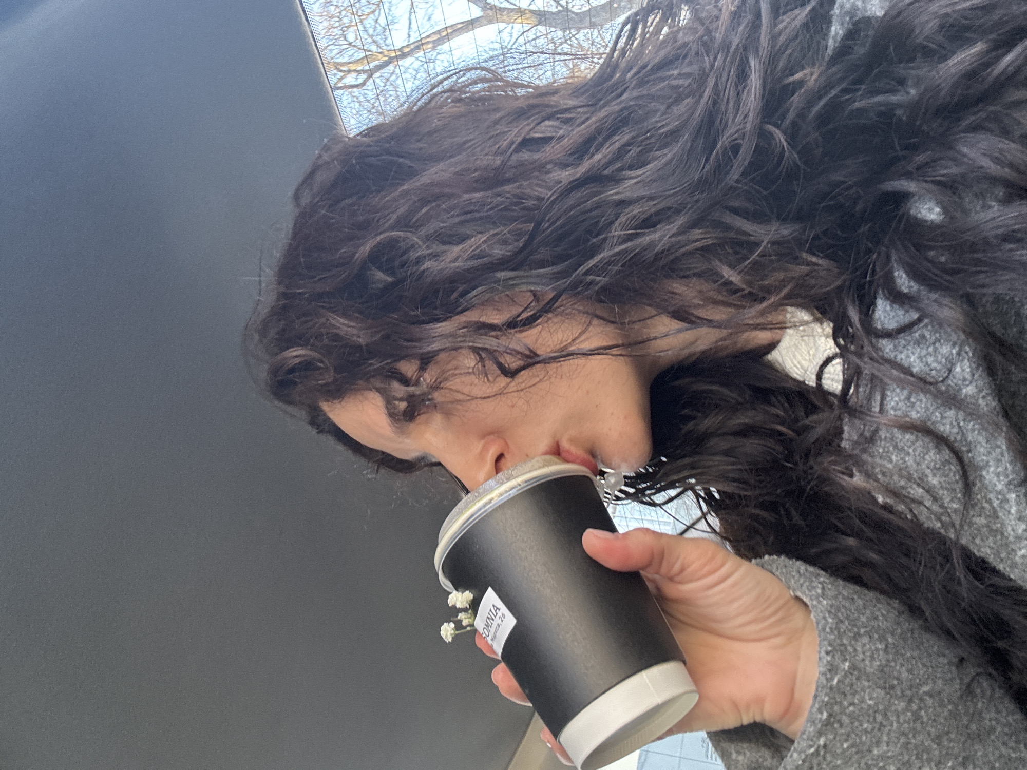 A female individual with long, dark, curly hair is depicted in profile, facing right, inside what appears to be a vehicle. She is wearing a grey textured outer garment over a lighter-colored collar. Her right hand holds a black paper cup with a white base and a clear plastic lid, from which she is actively drinking. The cup has a white label displaying "SOMNIA" and "Мира, 2Б". Small white flowers are inserted into the top of the cup, positioned above the label. The background visible through a window shows a clear blue sky and bare tree branches. The interior features a dark grey headliner. The address "Мира, 2Б" on the cup's label corresponds to "SOMNIA Coffee Shop" located on Mira Street 2B, in the city of Chaikovsky, Perm Krai, Russia.