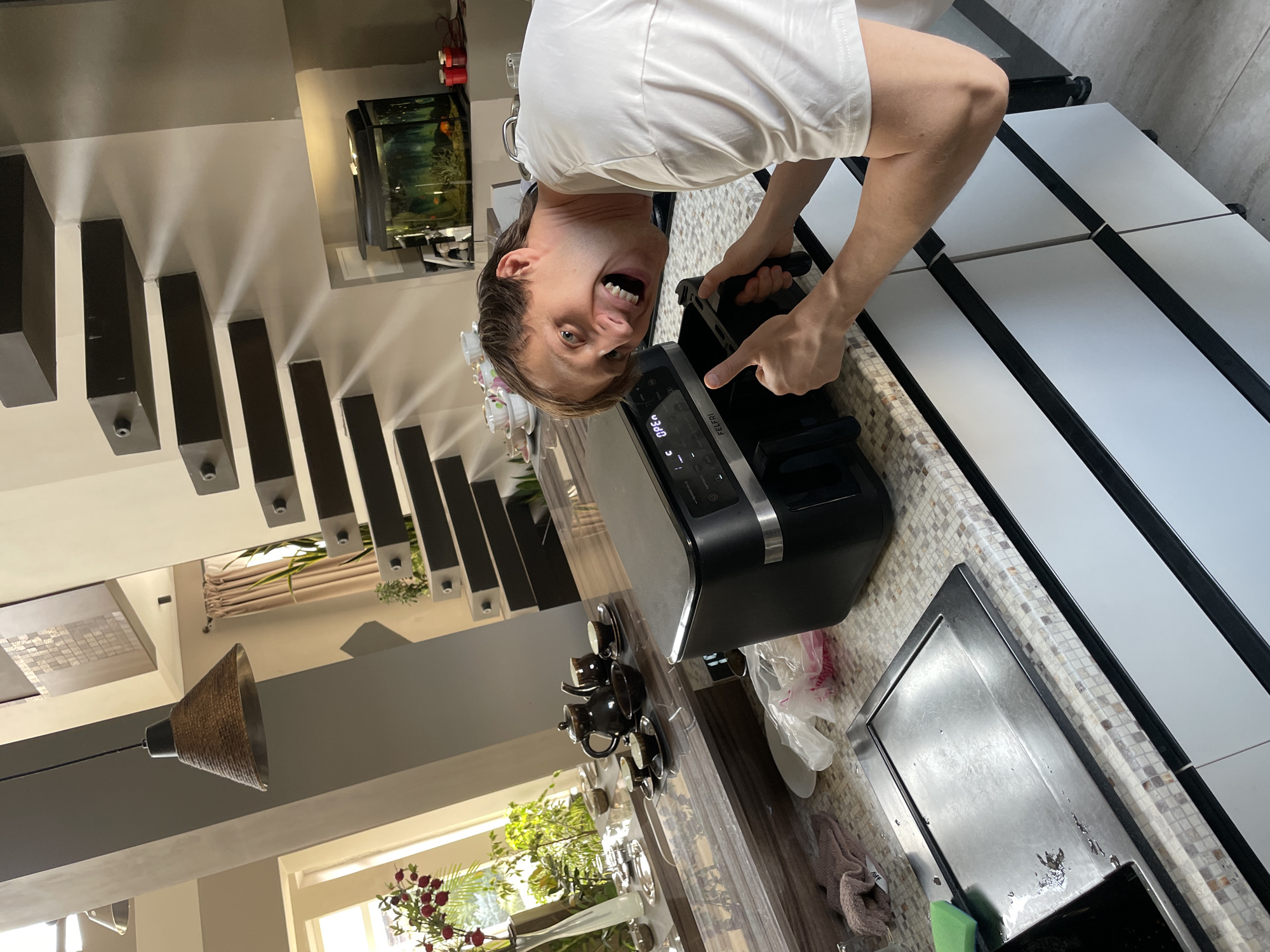 The image shows a person holding a black electronic device, likely an air fryer, in a kitchen setting. The person is facing the camera and has a wide, exaggerated smile. The kitchen appears modern with light-colored countertops and cabinets with black accents. A sink is present, and various kitchen items such as a teapot set, flowers in a vase, and cleaning supplies are visible. In the background, there are decorative stairs and a fish tank. The presence of Kurskaya Oblast, Russia can not be verified.