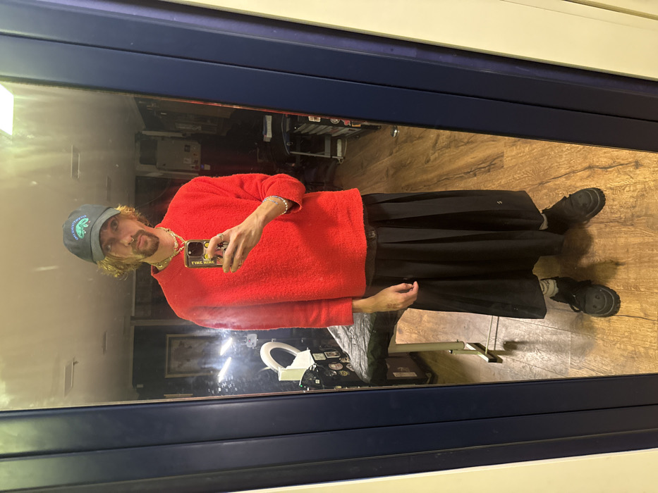 A person stands in front of a mirror, taking a selfie. The individual wears a red sweater, black skirt, black shoes, and a blue cap. Visible jewelry includes a necklace and bracelets. The background shows a room with various items, including a rolling chair, a light, and what appears to be a desk. The floor is made of wood. The mirror's frame has a blue border. The phone case has a word on it "FINE WINE".