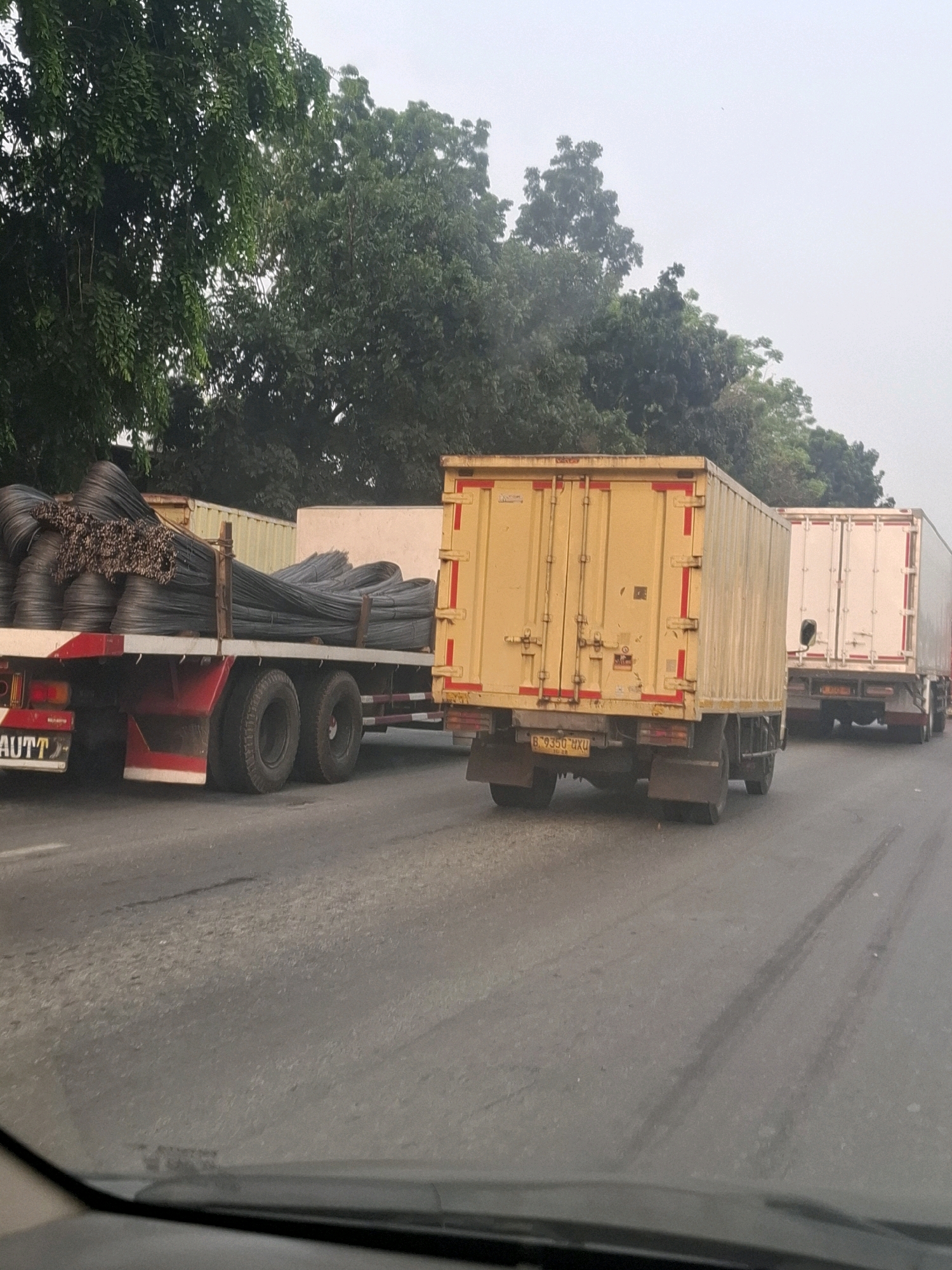 The image captures a scene on a paved road, likely in Bencongan, Indonesia, with a line of trucks visible. In the foreground, a flatbed truck is laden with bundles of dark metal rods, possibly rebar. The truck has dual rear wheels on each side and displays a red and white striped rear bumper. To its right, a yellow box truck is partially visible, followed by a white box truck. Trees with dense green foliage line the left side of the road, and the sky is overcast with a light haze. The road surface shows tire marks. The overall impression is a commercial transportation scene on a roadway.