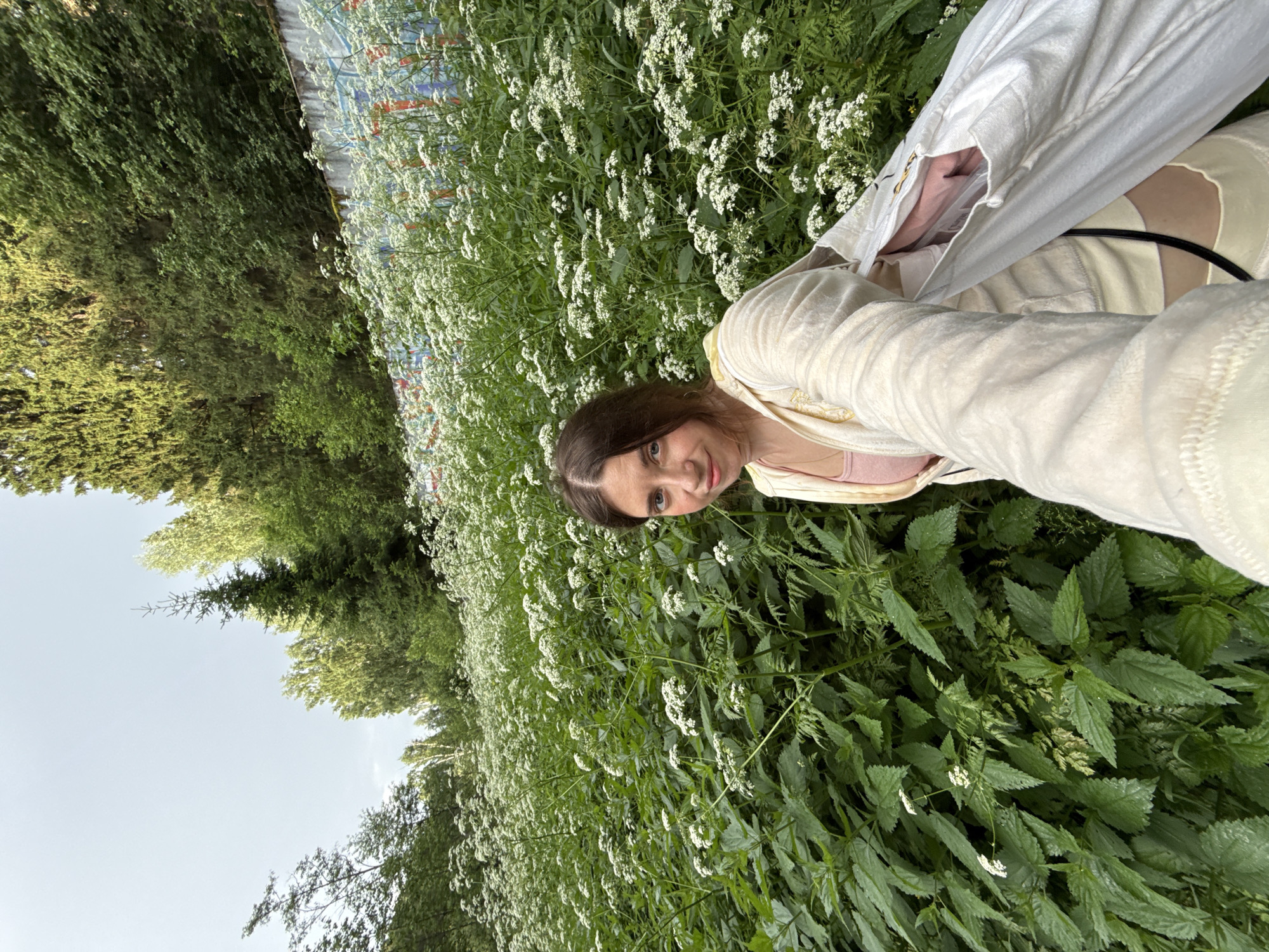 A woman is standing in a dense field of white wildflowers and green plants, looking at the camera. She has brown hair and is wearing a cream-colored zip-up hoodie over a pink tank top and yellow shorts. She is holding a white bag in her right arm. Behind her, there is a concrete wall with graffiti on it, and beyond that, a hill covered in trees. The sky is overcast.
