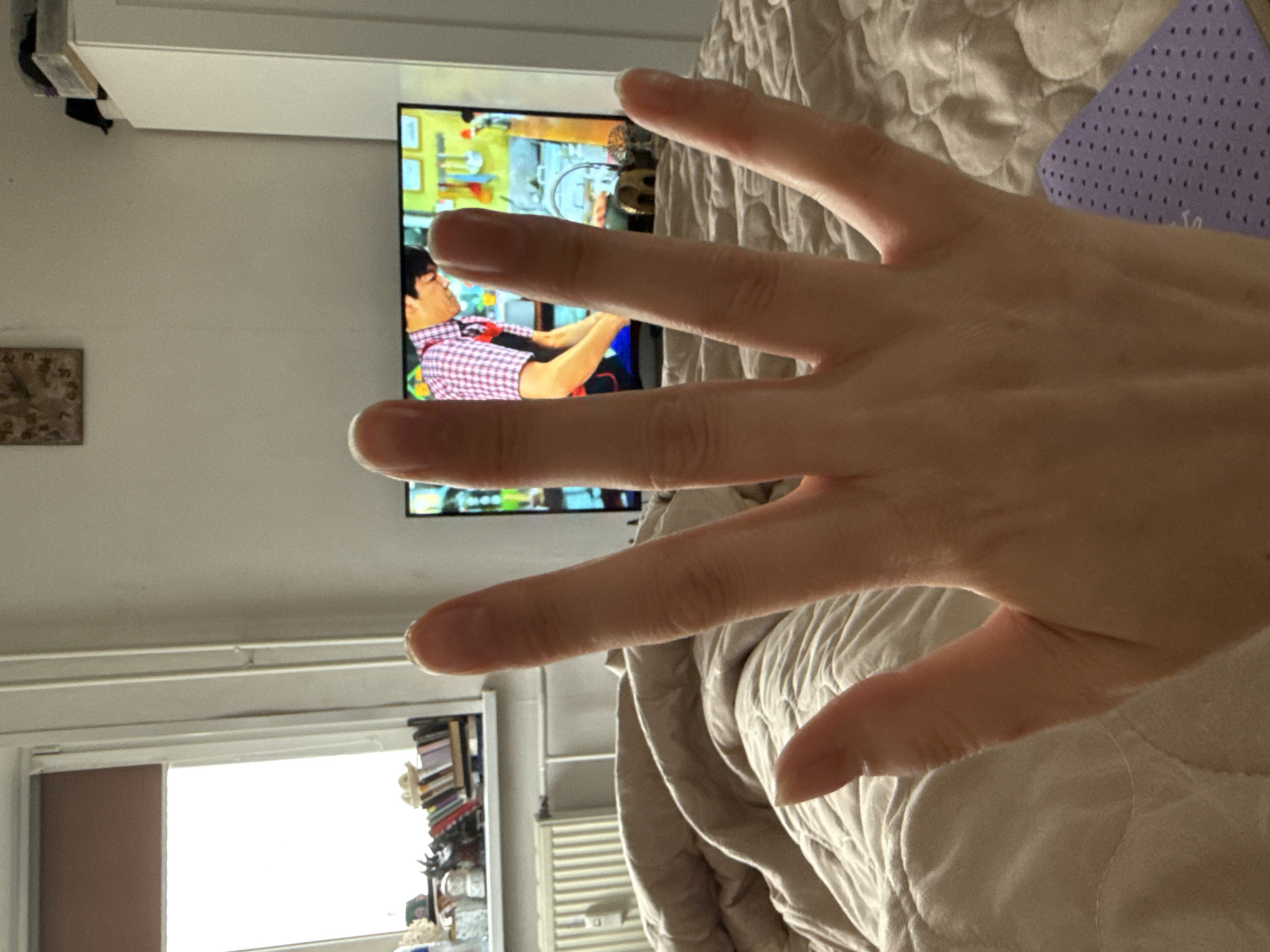 A hand is displayed in the foreground, partially obscuring a television screen showing a man in a red and white checkered shirt. The background features a window with a view of what appears to be an urban setting, possibly a residential or commercial area. The scene is indoors and seems to be in a bedroom based on the presence of a bed with a quilted comforter. The image does not contain information to determine the location in the city of Izhevsk, Russia.