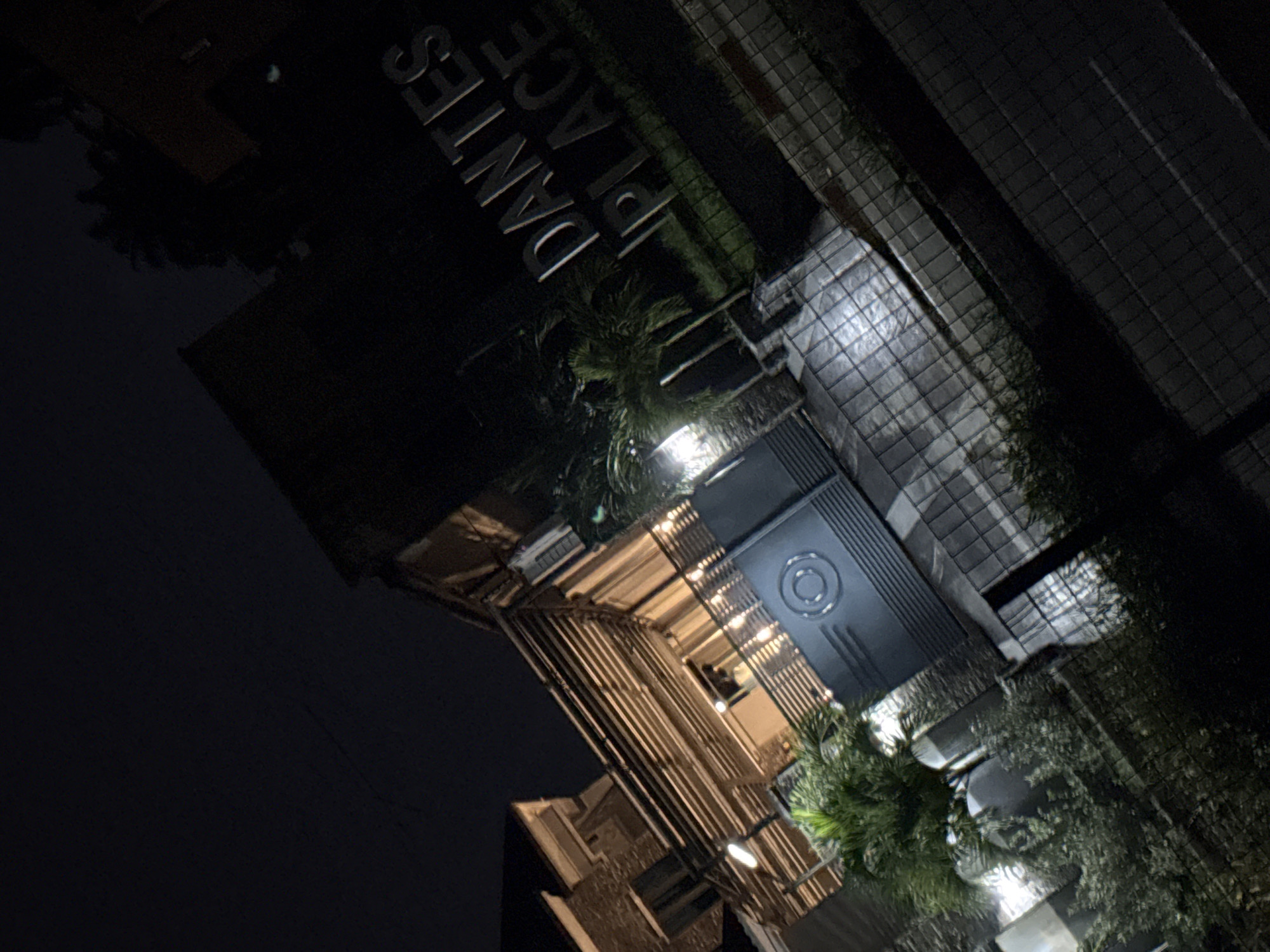 The image depicts the exterior of a building at night in Uyo, Nigeria. A sign with the words "DANTES PLACE" is prominently displayed in large, metallic letters. The building features a modern architectural style with an open, covered walkway constructed of horizontal wooden slats. A dark, imposing gate with a circular emblem is visible in the foreground, suggesting a secure entrance. Small palm trees and other foliage are planted around the entrance. The ground is paved with dark tiles or stones, and appears wet, possibly from rain. The overall lighting is dim, with some artificial lights illuminating the building and the entrance. The scene is angled, giving a dynamic perspective of the location.