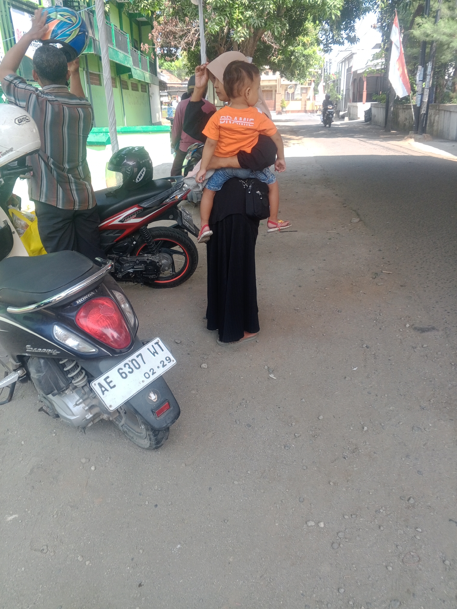 The image depicts an outdoor scene, likely in Banyudono, Indonesia, characterized by a gravelly road surface and a building in the background. In the foreground, a dark blue Honda scooter is parked, its rear license plate clearly visible and reading "AE 6307 WT". To the left, a red and black motorcycle is also parked. A man stands to the left, holding a multi-colored helmet above his head. A woman, dressed in a long black skirt and a light-colored headscarf, is carrying a young child on her hip. The child is wearing an orange t-shirt. The overall atmosphere suggests a casual, everyday moment in a semi-urban or roadside setting.