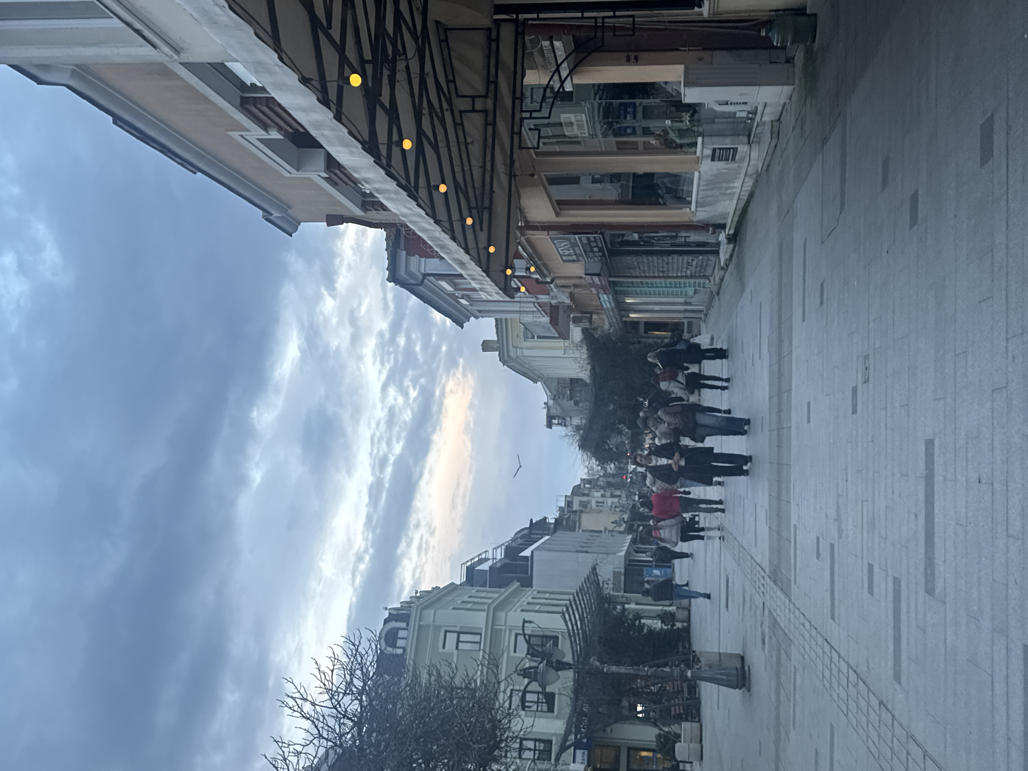 The image captures a street scene in Burgas, Bulgaria. The view is from a street-level perspective, looking down a pedestrianized street flanked by buildings on both sides. A group of people is walking in the same direction on the right side of the street. Overhead, decorative lighting is strung between the buildings. The sky is overcast with clouds. A bird is flying high overhead. The buildings are various colors and designs. The street is paved with large rectangular stones.