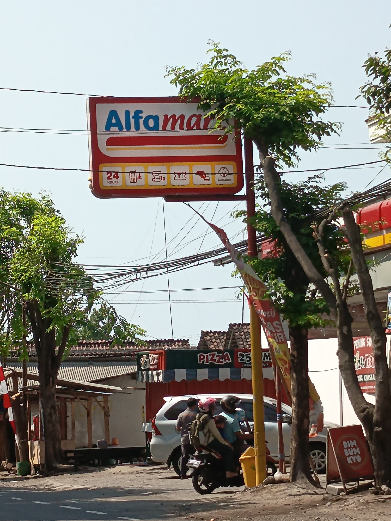 The image depicts a street scene in Kandangjati Kulon, Indonesia. In the foreground, a paved road with white dashed lines is visible. To the right, a red A-frame sign displays "DIM SUM KYO" in white lettering. Several trees with dark trunks and green foliage are present. In the mid-ground, a white car is parked. On the left side, a group of people on a motorcycle are riding past. The motorcycle has a license plate that reads "S 1821 XA". Behind the car and motorcycle, a low-rise building with a corrugated metal roof is visible, along with a smaller wooden stall. Further back, another building with a red and white striped awning has a sign that reads "PIZZA". Above the buildings, a prominent red and white sign for "Alfamart" is mounted on a pole, with smaller icons indicating services like "24 HOURS", a phone, an "Rp" symbol for currency, and another service icon. Several electrical wires are strung across the sky. The sky is clear and bright.