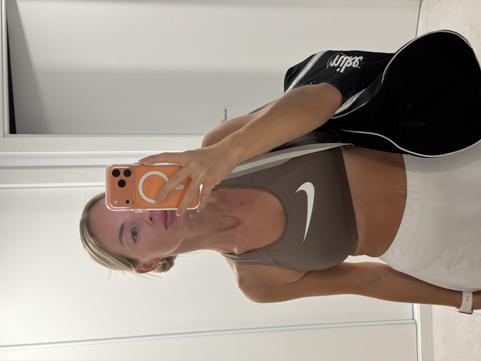 A fair-skinned woman with blonde hair pulled back in a bun takes a mirror selfie in an indoor environment, possibly a bathroom or dressing room. She is wearing a brown Nike sports bra and white athletic shorts. A black duffel bag with white trim and lettering is slung over her left shoulder. Her right hand is holding an orange smartphone, which is obscuring part of her face. The background consists of plain, light-colored walls and a doorframe. The scene is lit with artificial light. The location is not discernible from the visual information provided.