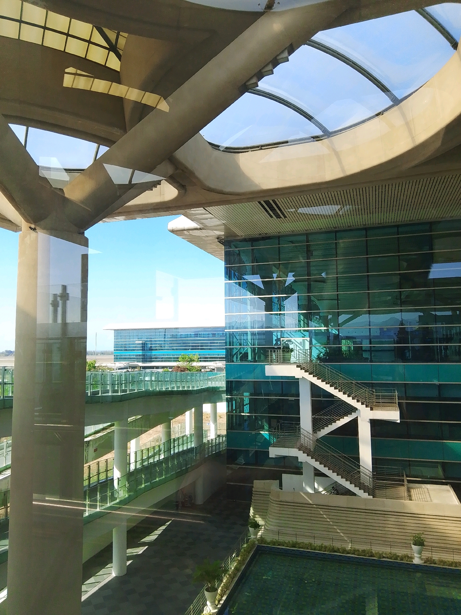 The image shows an exterior view of the Yogyakarta International Airport (YIA) in Palihan, Kulon Progo Regency, Special Region of Yogyakarta, Indonesia. The airport features modern architecture with extensive use of glass and concrete.
The foreground shows a large, partially reflective concrete pillar and structural elements of the airport's roof and walkways. A bright blue sky with some wispy clouds is visible through the translucent sections of the roof.
In the midground, a multi-story building with a striking teal-colored glass facade dominates the view. A prominent white outdoor staircase with metal railings ascends alongside the glass building. Reflections of the sky and surrounding structures are visible on the glass.
Further back, another long, low-rise building with a glass facade is visible, suggesting terminal or operational areas. Greenery, including trees, is interspersed around the buildings.
The lower part of the image reveals a paved walkway with patterned tiles and linear shadows cast by the overhead structures, indicating a sunny day. A shallow, tiled pool with a dark green surface and some floating debris is visible in the bottom right corner.
There are no people clearly visible in the image. The scene depicts a well-maintained, modern airport environment.
