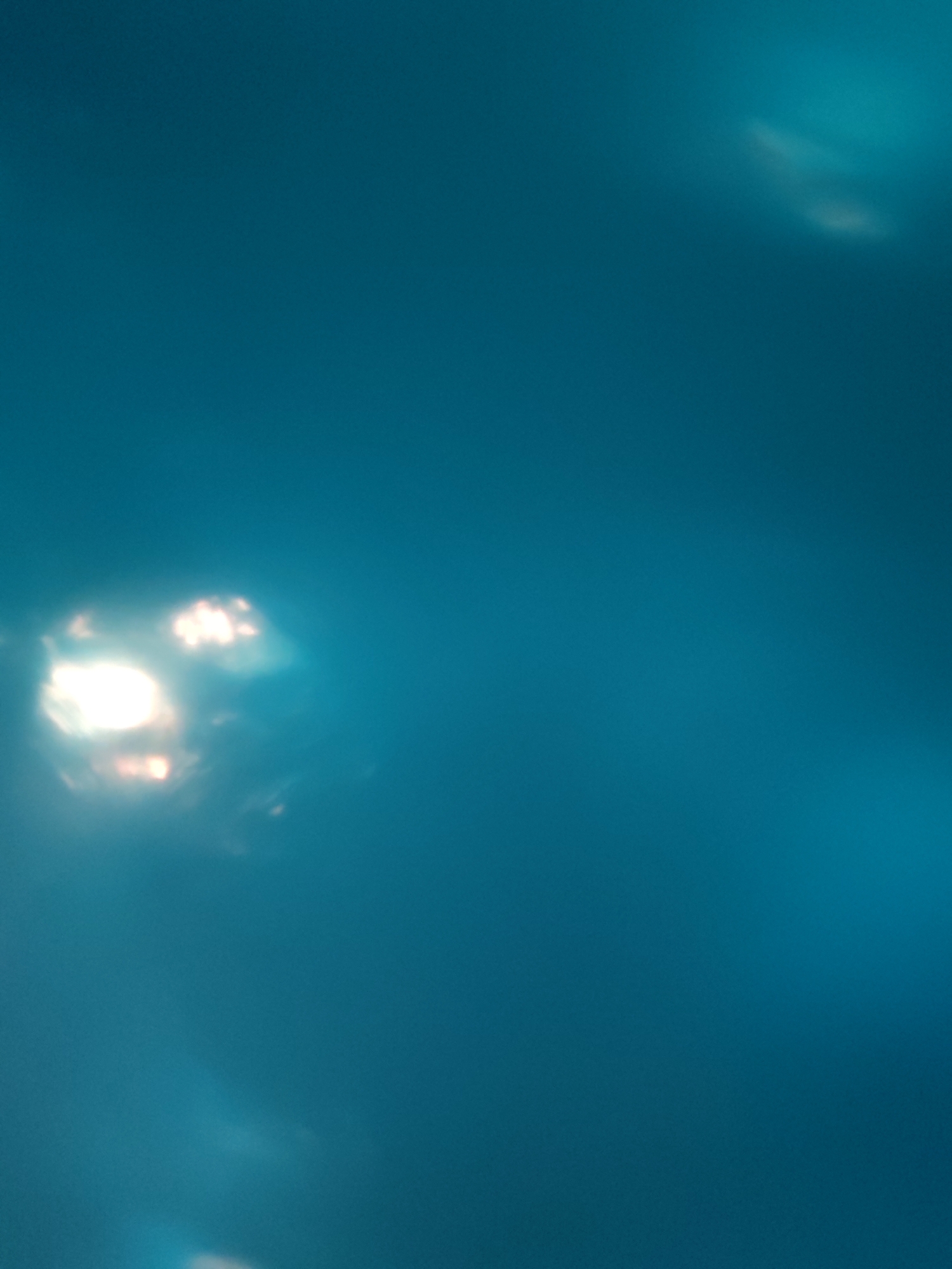 The image shows a blurred view, predominantly of a turquoise-blue color. In the lower left corner, there is a bright, unfocused patch of light, with some hints of orange/yellow within. There's another patch of light, fainter, at the top right. The overall impression is of an out-of-focus scene with light reflecting off a blue surface. Given the limitations of the image, providing details regarding the location in the city of Bol'shie Mkhi, Russia, is not possible.