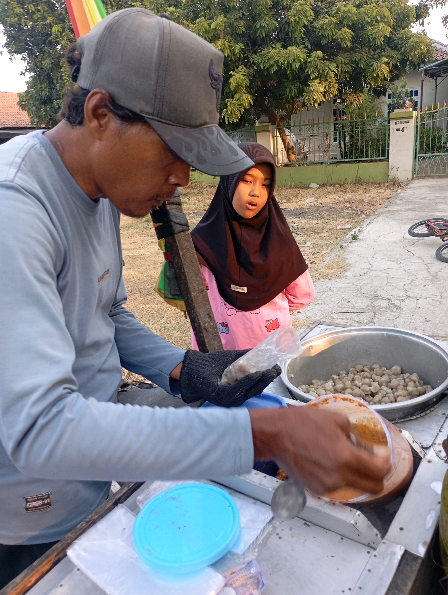 A man wearing a gray baseball cap and a light blue long-sleeved shirt is serving food from a cart. He is holding a clear plastic bag with food items and a spoon, and appears to be scooping sauce into a plastic container. Next to him, a young girl in a black hijab and pink shirt with cartoon characters is looking at the man. In the background, trees, a house with a white fence, and a street are visible. The scene is outdoors, likely on a street in Babelan, Indonesia, given the context provided.