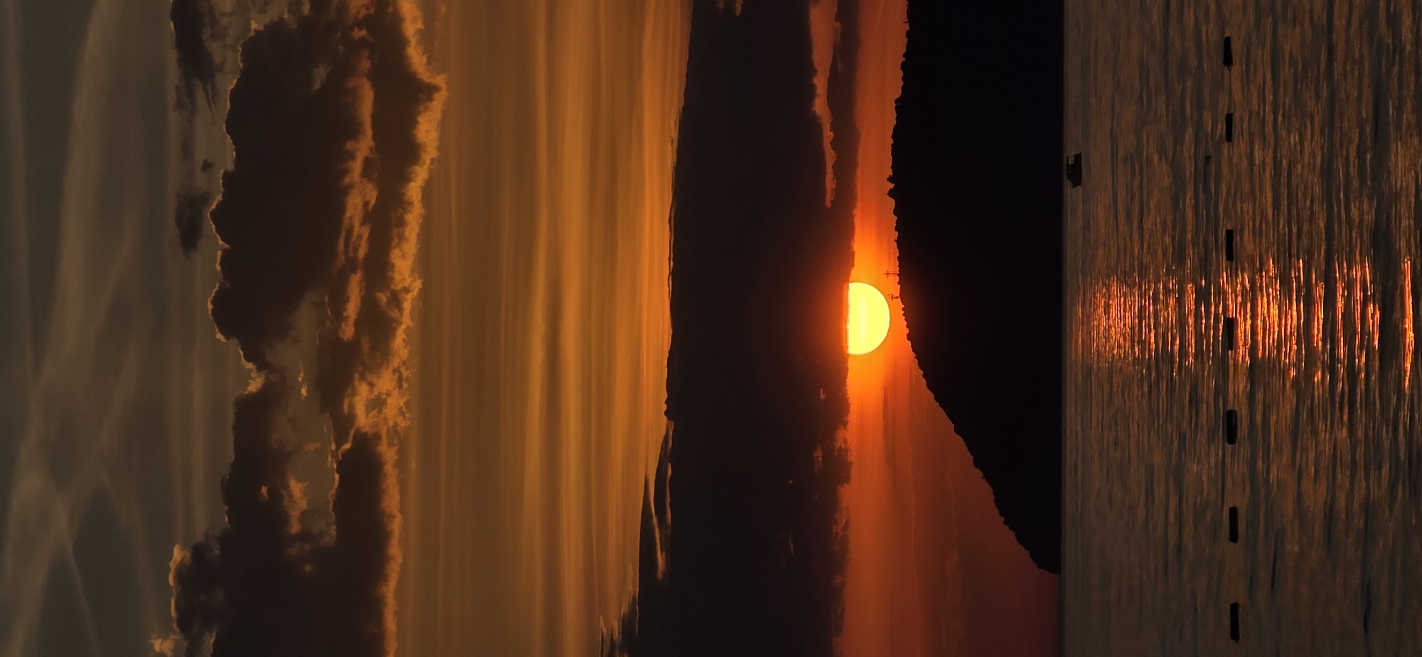 A rotated, long shot captures a sunset scene. The sky is a deep orange, filled with dark, textured clouds. A half-sun peeks from behind a black, silhouetted landmass. The sea reflects the orange light, with scattered, dark objects puncturing the surface. The location is the city of Bar, Montenegro.