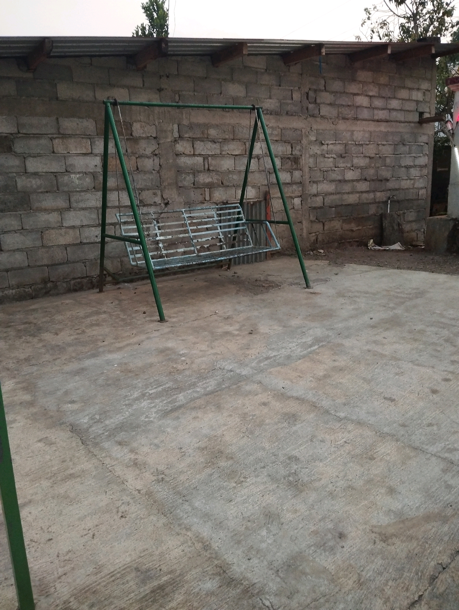 A green metal swing set with a white slatted seat is positioned on a concrete outdoor patio in front of a cinder block wall. The swing set is the primary object of focus. The wall appears to be part of a building with a corrugated metal roof. The ground is a rough, uneven concrete surface. The lighting suggests it is daytime, possibly late afternoon or early morning, with soft, diffused light. The environment is outdoors and appears to be a private yard or common area. The image does not contain any people or visible actions. There is no indication of the specific location within Salatiga, Indonesia.