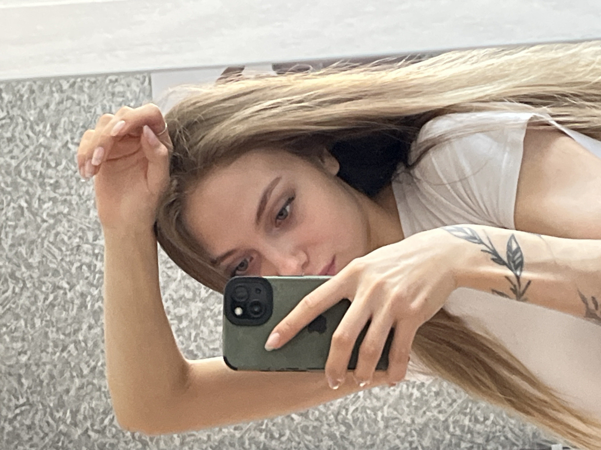 A woman is taking a selfie in front of a mirror. She is holding a green iPhone in her right hand, and her left arm is raised near her head. She has long blonde hair and is wearing a white t-shirt. There is a floral tattoo on her left wrist. The background appears to be a wall with a textured pattern. There is no information to suggest this photo was taken in the city of Фоки, Russia.