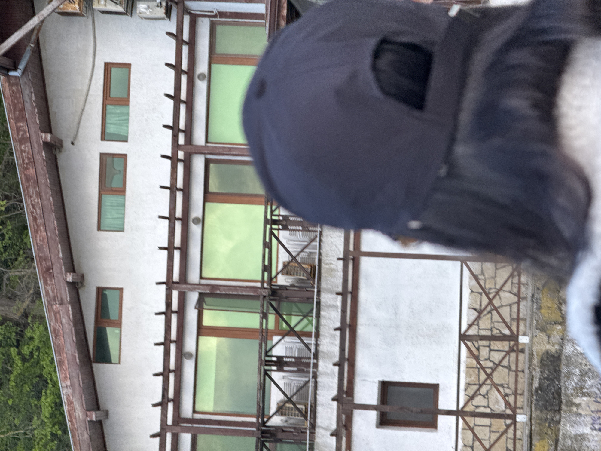 A building is seen with white walls and brown window frames. The windows reflect green hues. A black baseball cap is in the foreground, partially obscuring the view. The photo appears to have been taken from above, looking down at the building. The structure has a wooden framework extending from the wall. Vegetation, possibly trees, is visible on the left side of the image.