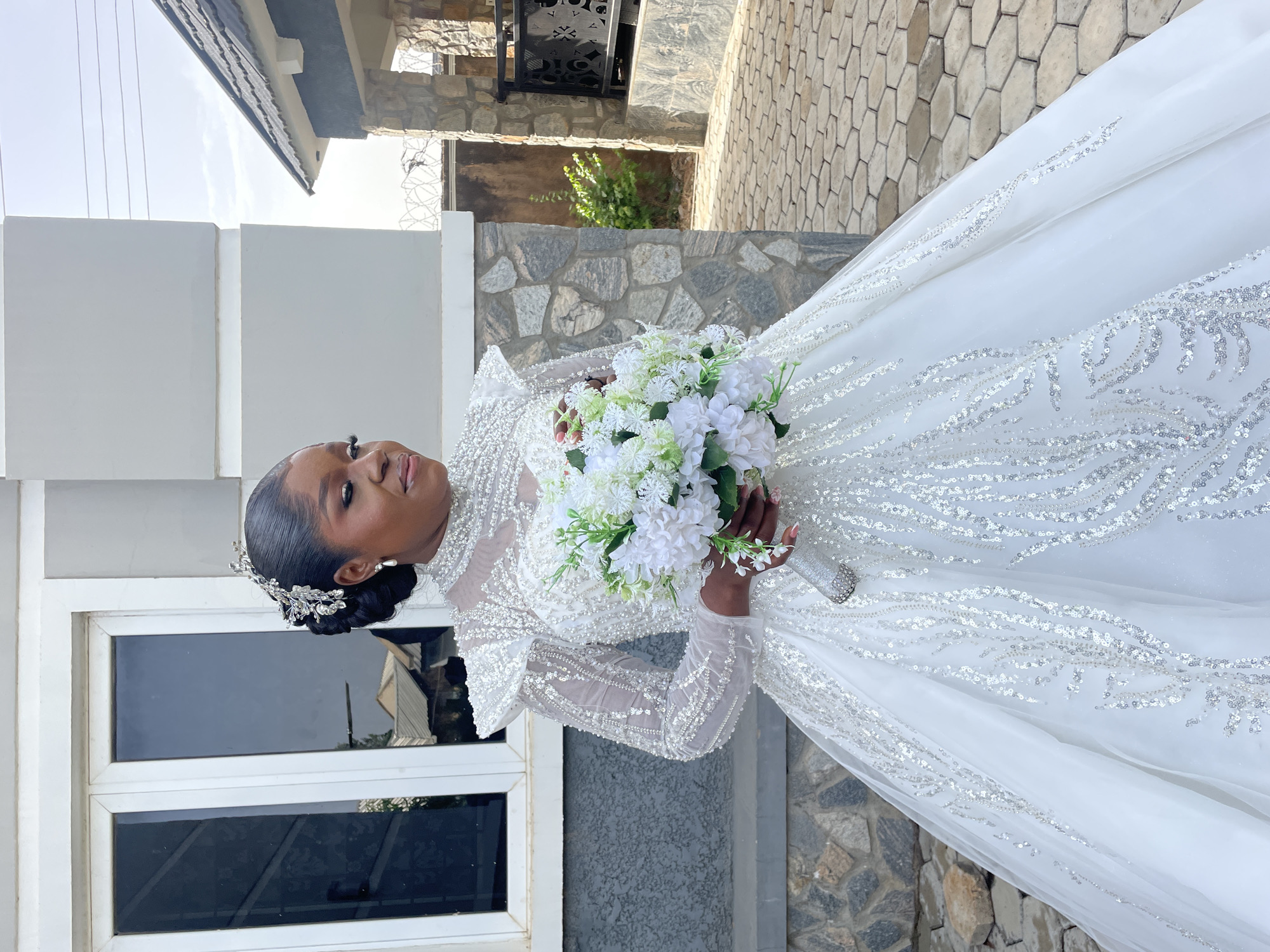 A woman in a long-sleeved, white and silver beaded wedding gown stands outside a building. She holds a white floral bouquet. Her hair is styled up with a jeweled headpiece. She looks to her right. The building behind her has a gray stucco exterior and a white-framed window. The ground and a portion of the building feature stone cladding. While the city of Bukuru, Nigeria is requested, this cannot be determined from the image.