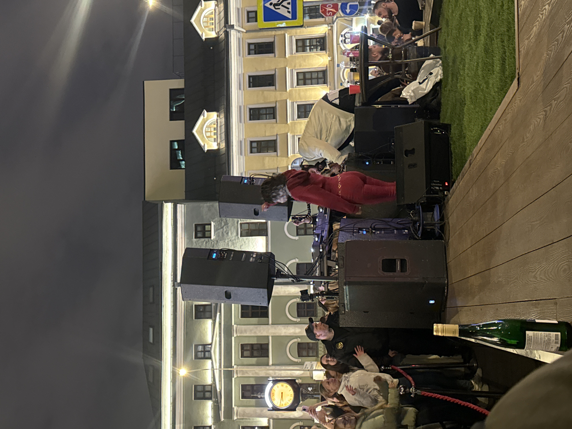The image shows an outdoor setting at night in Moscow, Russia, with a live music performance. A building with multiple windows and architectural details is visible in the background, along with a clock tower. On a raised platform covered with wooden planks, musicians perform behind speakers. A woman in a red jumpsuit is visible among the equipment. A small patch of artificial green turf is present to the right. In the foreground, a crowd of people is watching the performance. A bottle of wine lays on the wooden platform edge. Overhead, a street light illuminates the scene.