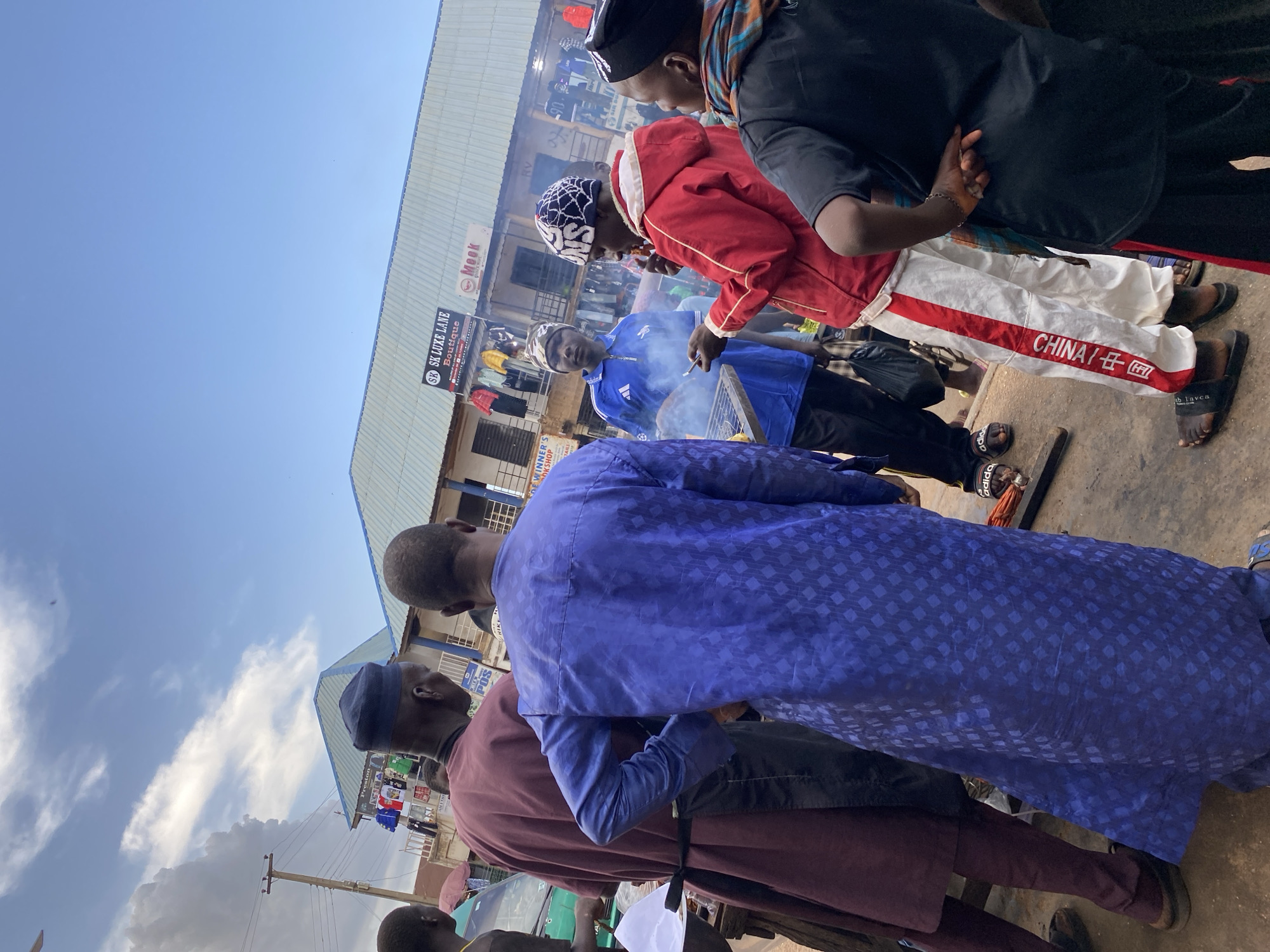 The image depicts a group of people gathered in Bukuru, Nigeria. A man in a red jacket and another in a blue shirt appear to be grilling food on a small barbecue. The group is standing outside a building with signs indicating various businesses, including "SK LUXE LANE Boutique" and "WINNER'S OKSHOP". Another sign reads "Mook".
One man is wearing trousers with "CHINA/ 中国" written on them. Several men are wearing traditional robes. The sky is clear and blue. A power pole is visible in the lower left corner of the image.
