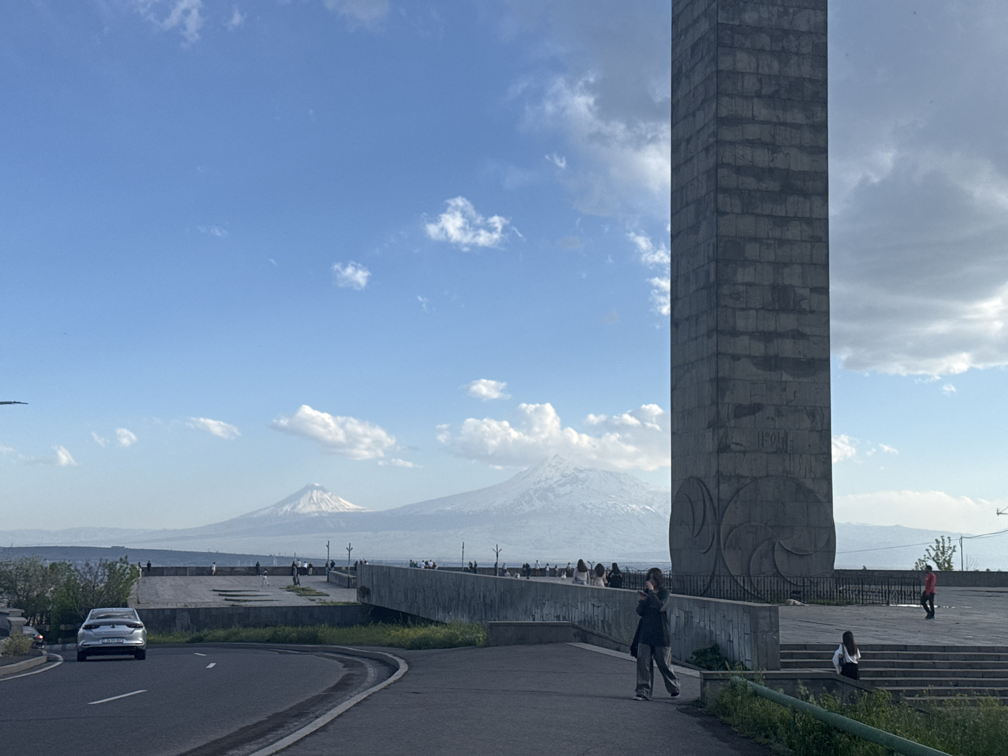 The image shows a scene in Yerevan, Armenia, with Mount Ararat visible in the background.
**Environment:** The setting is a public space near a road. The sky is partly cloudy with blue patches. The landscape includes distant mountains with snow-covered peaks.
**Objects:** A tall, rectangular, gray stone monument stands prominently on the right side of the image. A bridge or elevated platform connects the monument area with another viewing area to the left. A road curves in the foreground, with a silver car driving on it.
**People:** Several people are present in the scene. Some are standing on the platform or bridge, appearing to be enjoying the view. A woman is walking on the pavement near the base of the monument. One person is descending some stairs.
**Actions/Interactions:** People are walking, standing, and observing the view. The car is moving on the road.