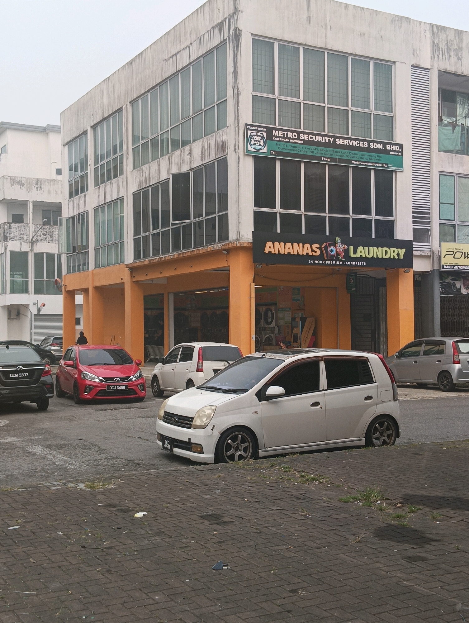 The image shows a commercial building in Kuching, Malaysia, with multiple storefronts. A prominent sign on the building reads "METRO SECURITY SERVICES SDN. BHD." and provides an address in the Commercial Centre, off Jalan Lapar, Samarahan. Another sign above a laundrette reads "ANANAS LAUNDRY 24-HOUR PREMIUM LAUNDRETTE." Several cars are parked in front of the building, including a black sedan with license plate OCM 9307, a red hatchback with license plate OCJ 6456, and a white hatchback with license plate OCC 4439. The building appears to be slightly aged, with some discoloration on its exterior. The sky is overcast, and the environment suggests a commercial or mixed-use urban area. There is one person visible in the image, standing near the red car.