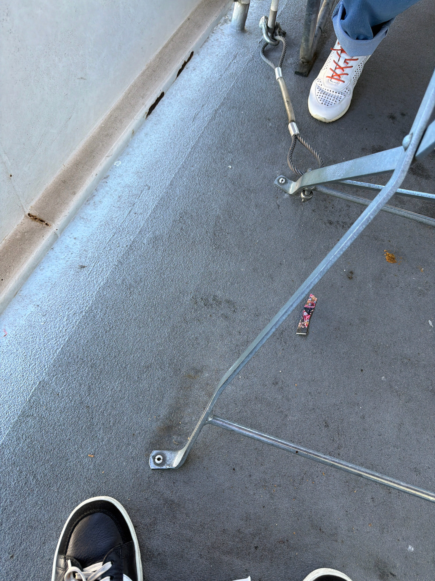 An outdoor scene on a gray, textured surface with a lighter colored concrete edge. A person wearing blue pants and white sneakers with orange laces stands on the right side of the frame, their left leg bent. Below them, to the left, is a close-up of another person's feet wearing black sneakers with white laces. In the foreground, a metal structure with thin legs and a diagonal bar is visible. A small, colorful fabric band with a floral pattern lies on the gray surface near the metal structure. The scene appears to be a walkway or deck, possibly near a body of water given the typical ambiance of the Romanshorn promenade.