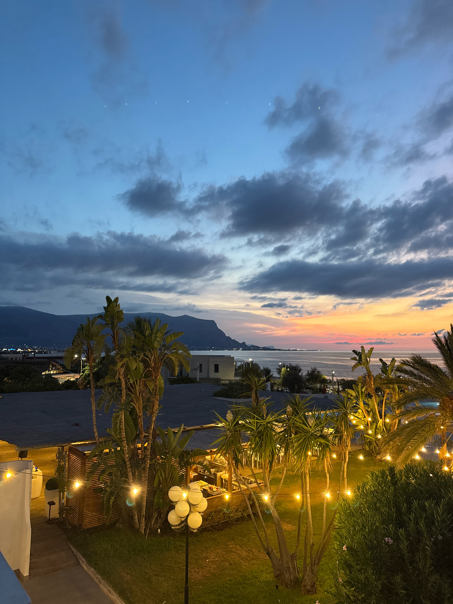 The image captures a dusk scene in Isola delle Femmine, Italy, overlooking the sea and mountains. The sky is a gradient of deep blue at the top, transitioning to lighter blues and streaks of orange and pink near the horizon, indicating the last moments of sunset. Several thin, white objects are visible in the upper portion of the sky, appearing as faint lights or reflections. Dark clouds are scattered across the sky, adding texture and depth.
In the foreground, lush green grass is illuminated by string lights and a multi-globe lamp, creating a warm, inviting atmosphere. Several palm trees and other tropical foliage are present, some adorned with hanging string lights. A wooden structure with a slatted roof, possibly an outdoor dining area or bar, is visible in the middle ground, with lights illuminating the interior. Beyond this, the calm sea stretches out to the horizon. In the distance, the coastline is dotted with lights from buildings, and a range of dark, imposing mountains forms a backdrop against the colorful sky. A white building is also present in the middle ground. The overall scene suggests a serene evening setting by the Mediterranean. There are no people or animals visible in the image.